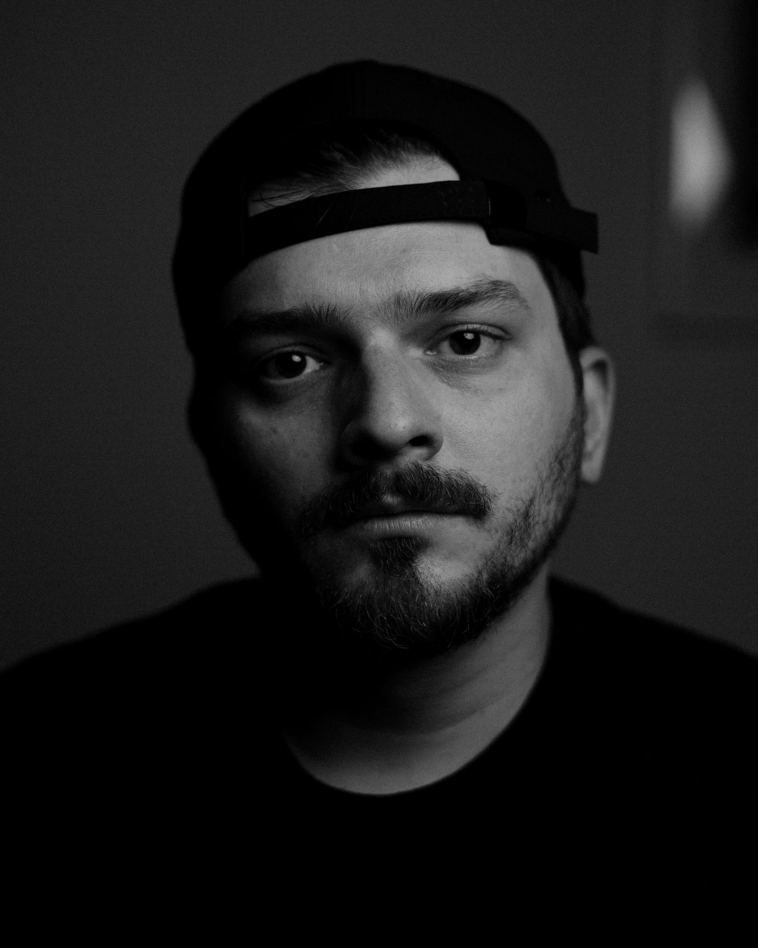 Black and white portrait of Jenya Mironov in a backwards cap and dark tee, lit from one side against a dark wall, looking straight into the camera