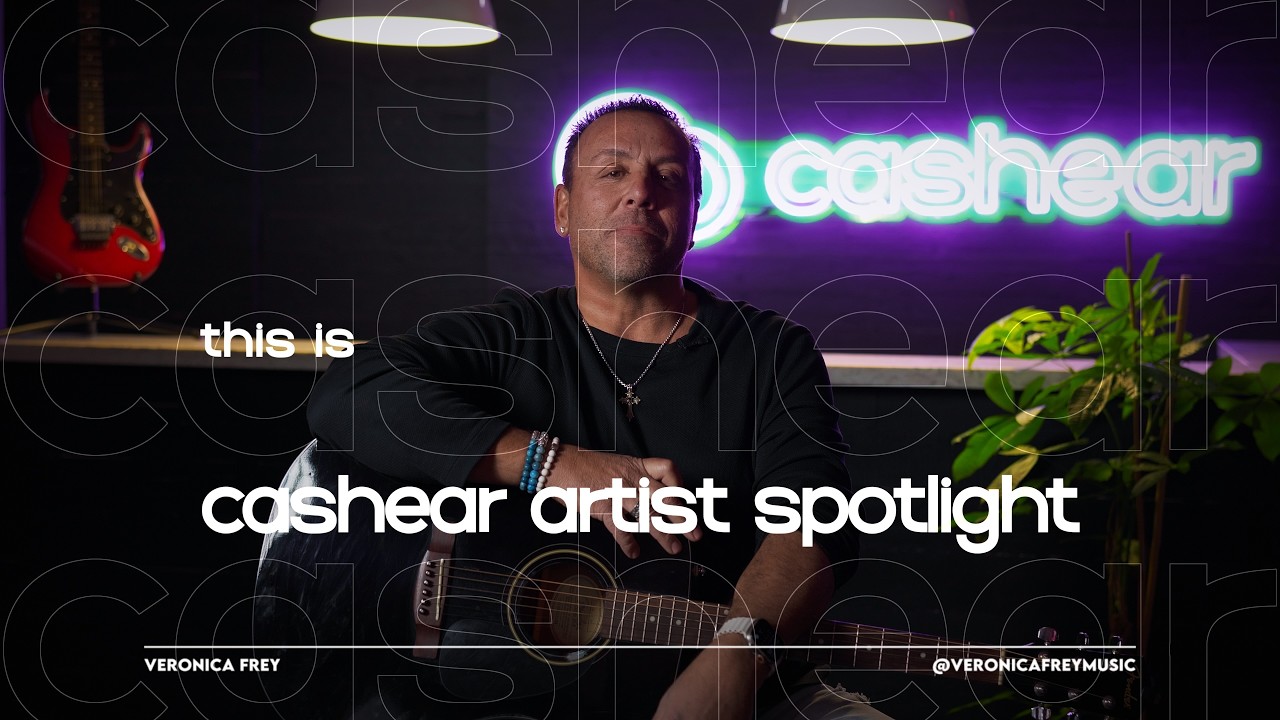 A man in a black tee and silver chain holds an acoustic guitar on the Artist Spotlight set, the neon cashear sign lit behind him