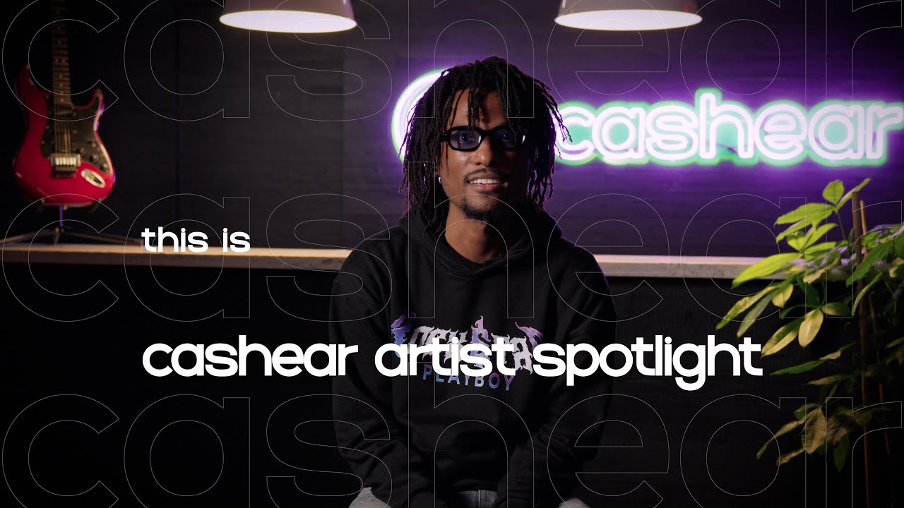 FritzThaProducer in glasses and a black hoodie on the Artist Spotlight set, the neon cashear sign behind him