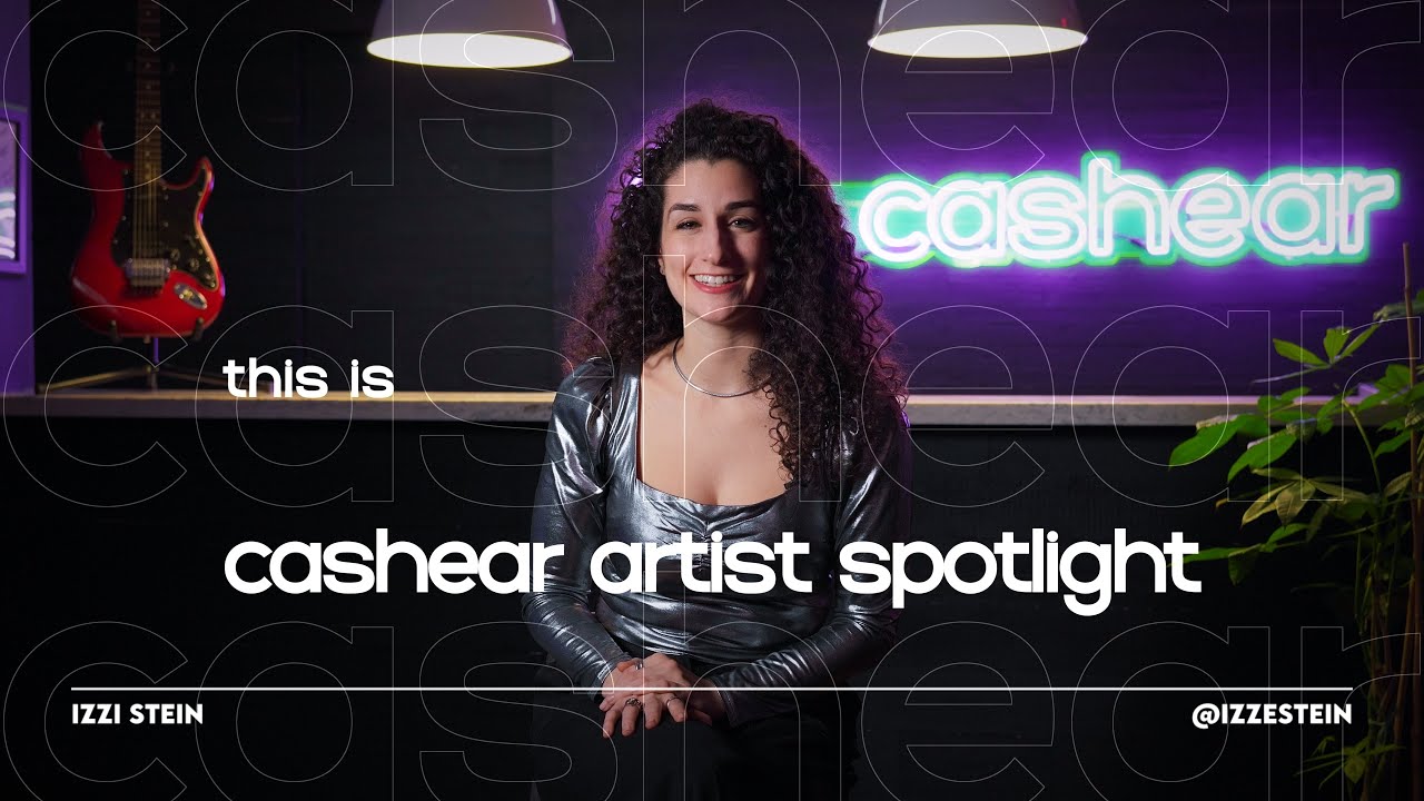 Izzy Stein smiling in a metallic silver top on the Artist Spotlight set, dark curls past her shoulders