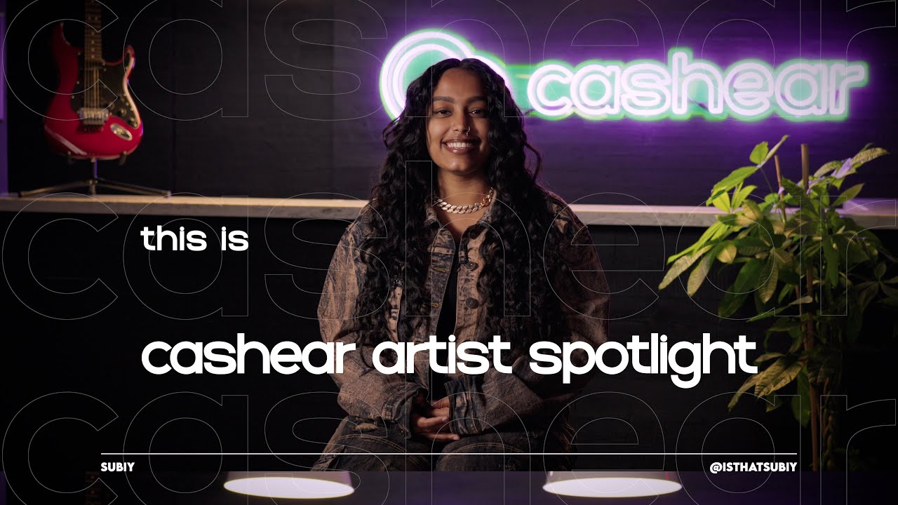 Subiy smiling in a patterned jacket and gold chain on the Artist Spotlight set under the neon cashear sign