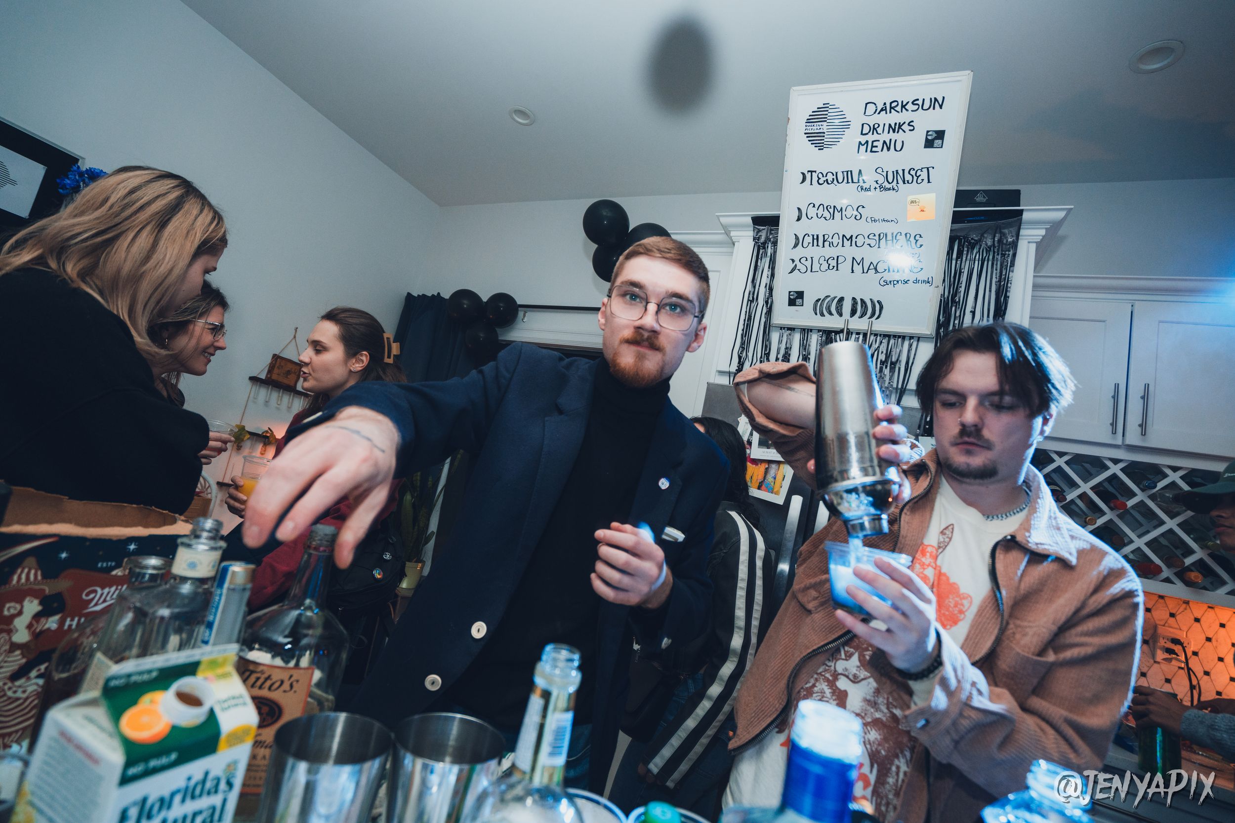 Bartender in a dark blazer reaches across a crowded cocktail counter below a handwritten drinks menu at a private party