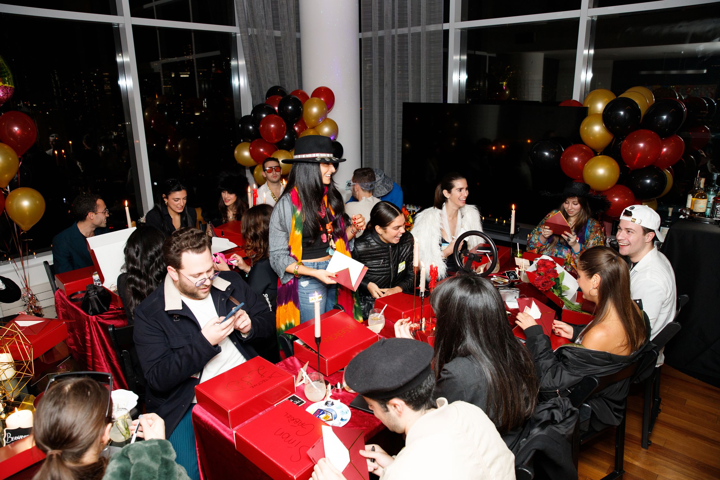 Guests at tables covered in red cloth with red gift boxes and lit candles, black and gold balloon columns behind them and the city night in the windows