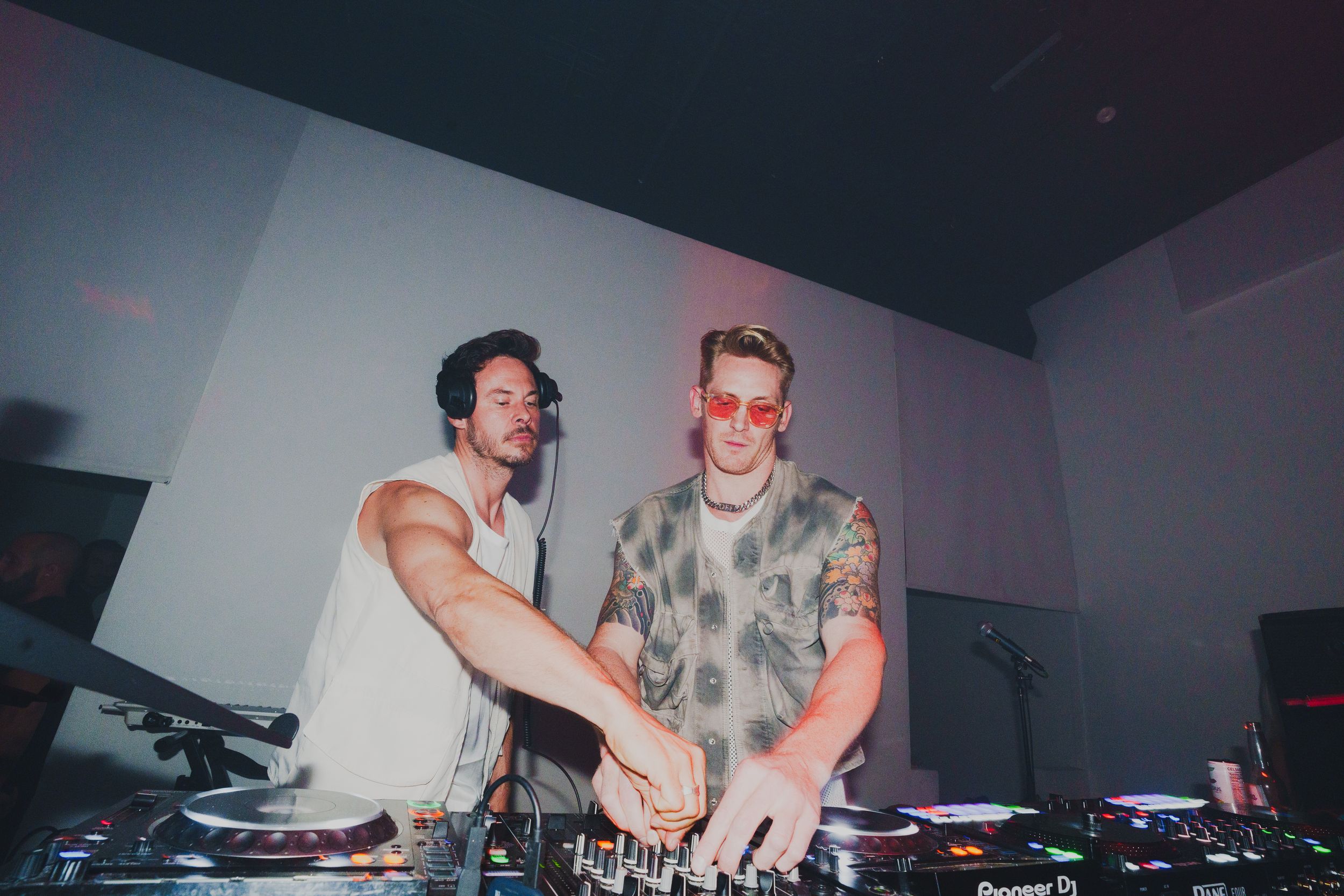 Two DJs behind a mixer with their arms over the decks, lit from the side in a loft with white walls