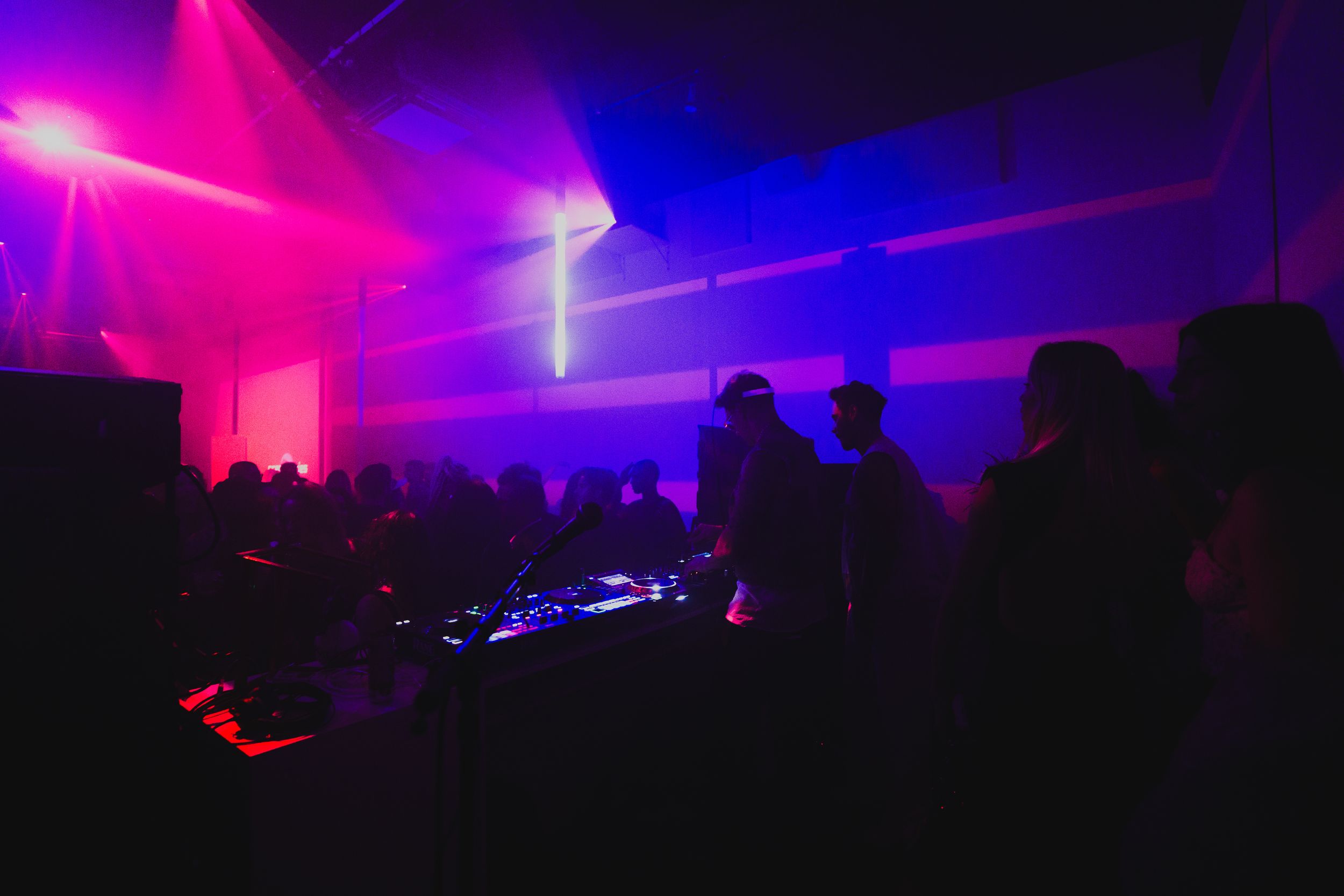 A crowd in violet and blue haze under crossing light beams, facing a DJ booth at the back of the room