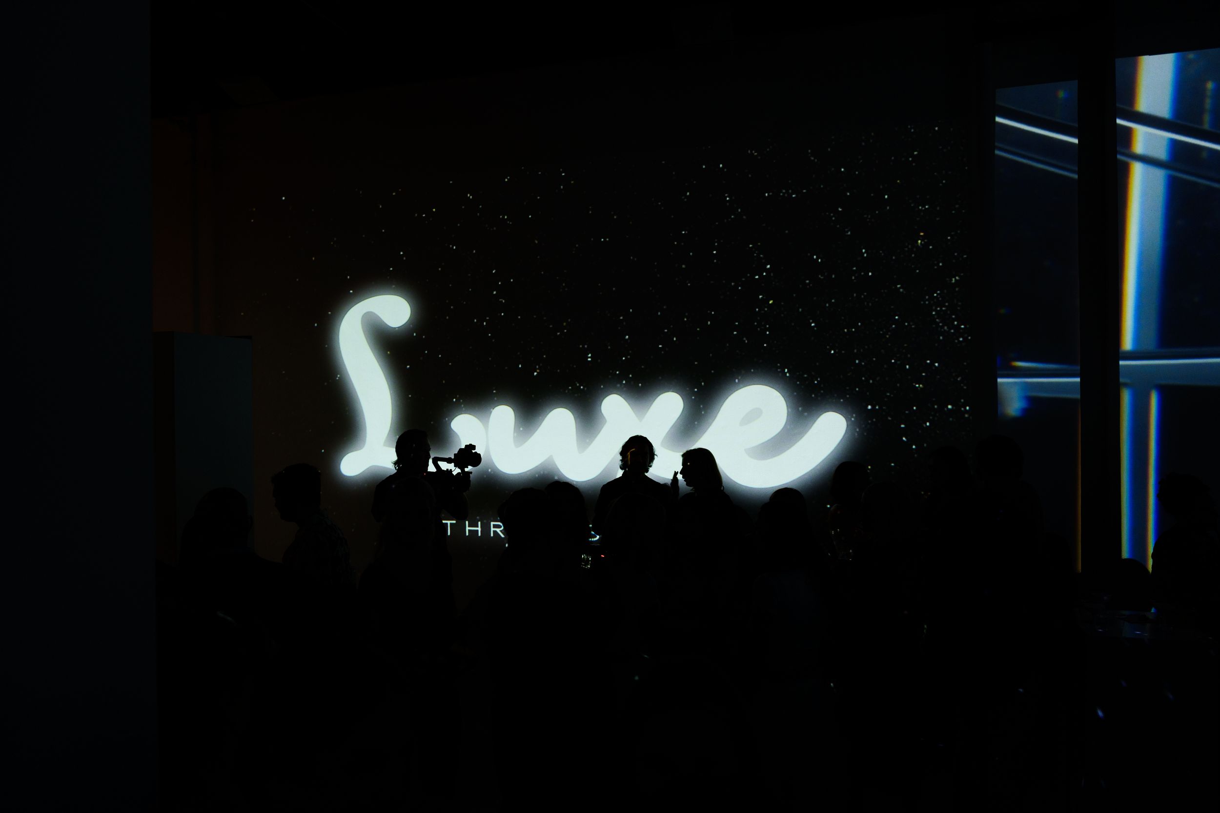 A white neon script sign glowing on a dark wall above a small crowd