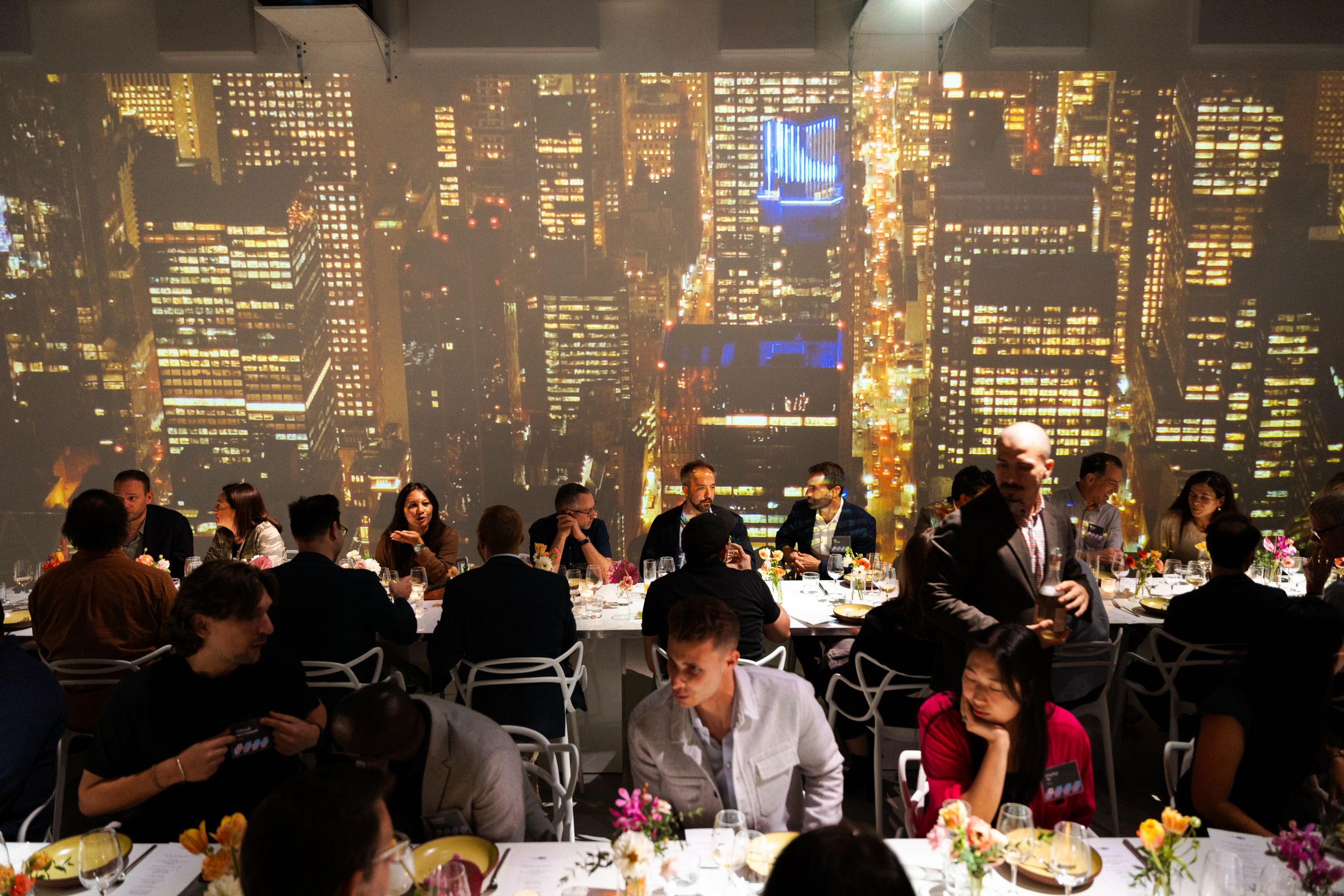 Guests at long dinner tables on a high floor, the lit towers of the city filling the windows behind
