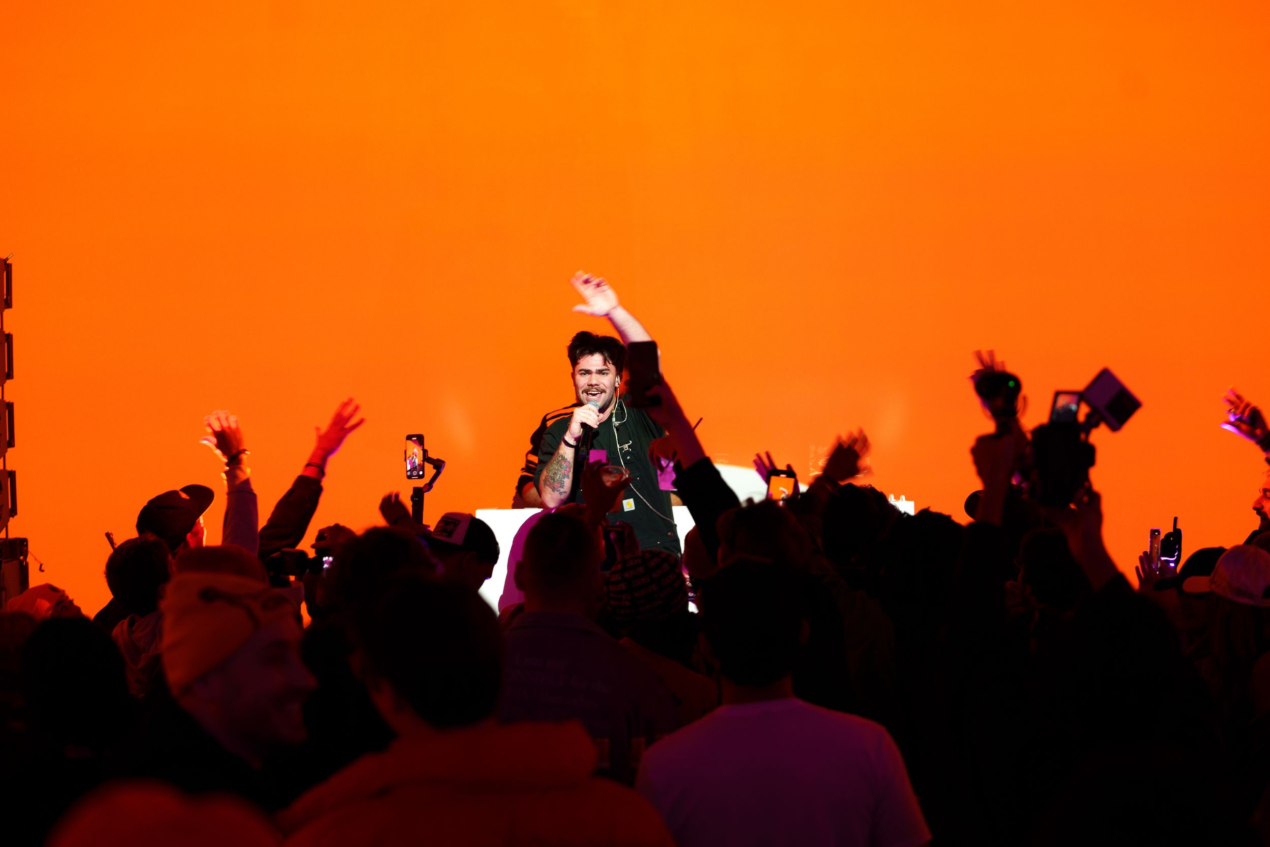 A performer with a microphone raising one arm on a lit riser against a wall of orange light, above a silhouetted crowd holding phones up