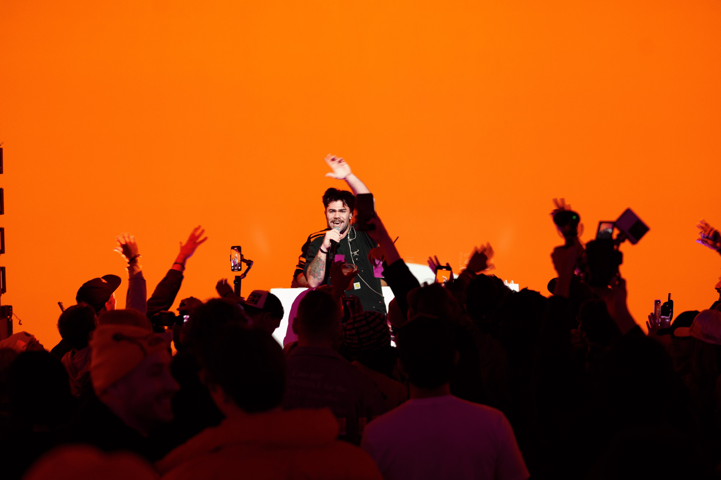 Performer with a microphone lit against a bright orange backdrop, one arm raised, above a silhouetted crowd holding up phones