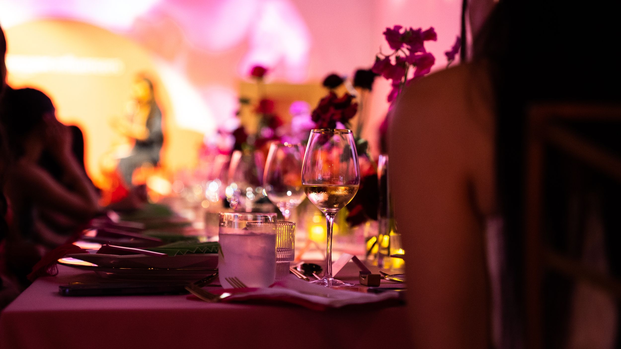 Candlelit dinner tables set with glassware and flowers, a guest passing through the warm pink light