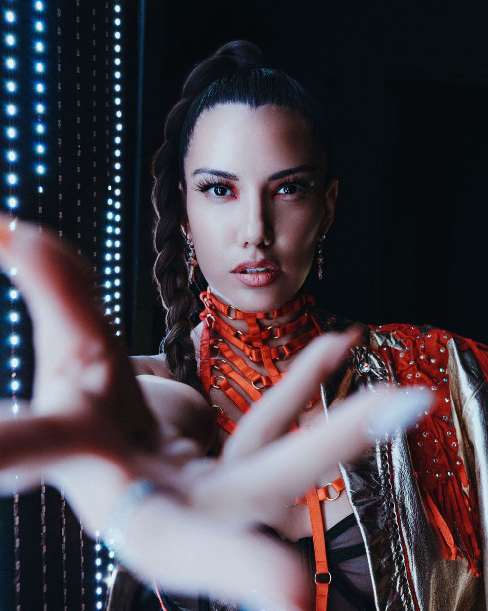 Fashion portrait of a model in an orange strap harness and a red embellished jacket reaching toward the camera, lit against vertical LED light strips