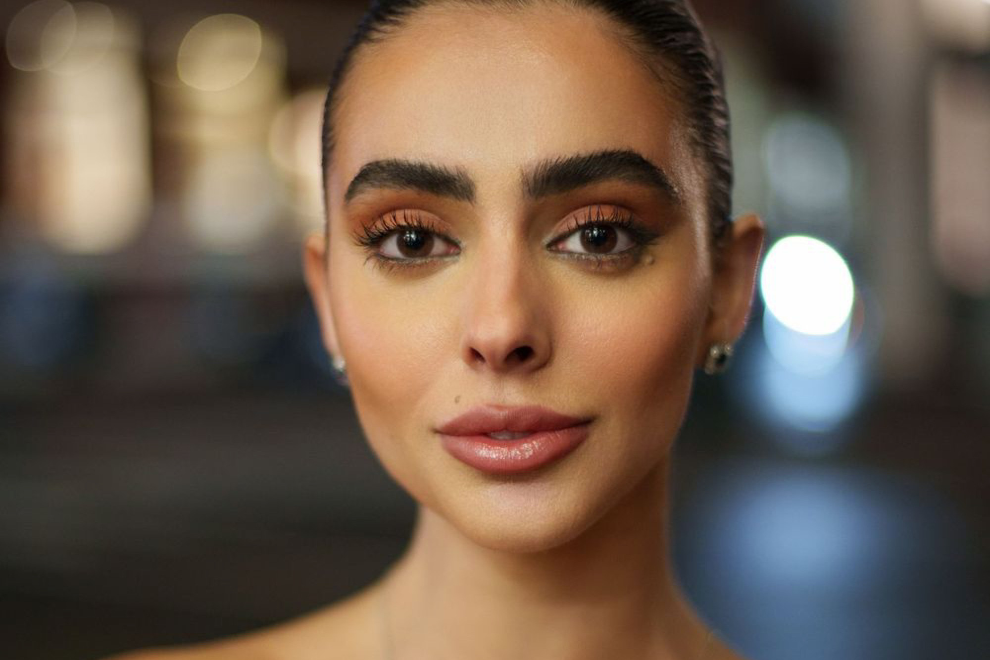 Beauty portrait of a woman with slicked back hair, defined brows and warm evening makeup, lit against blurred night lights