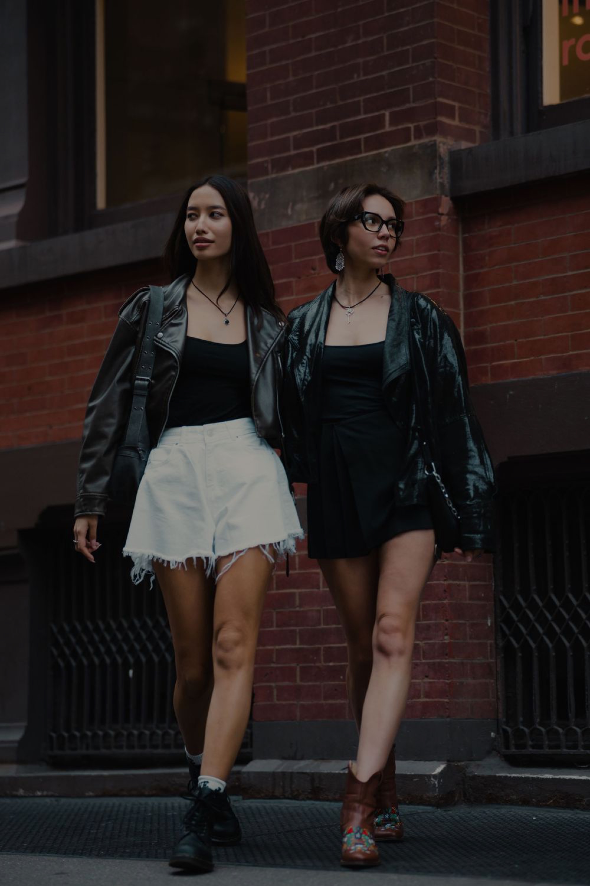 Two models in leather jackets and contrasting outfits posing against a SoHo brick building