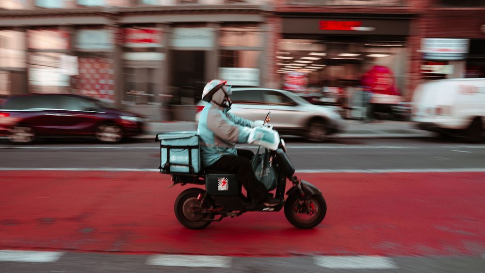 A delivery rider on a motorcycle crossing a stretch of street painted red, past parked cars