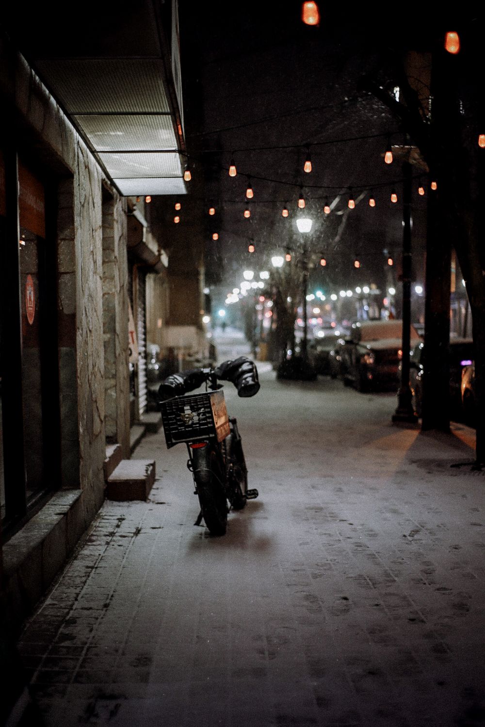 A downtown block covered in snow at night, motorcycles parked under a string of bulbs along a scaffold
