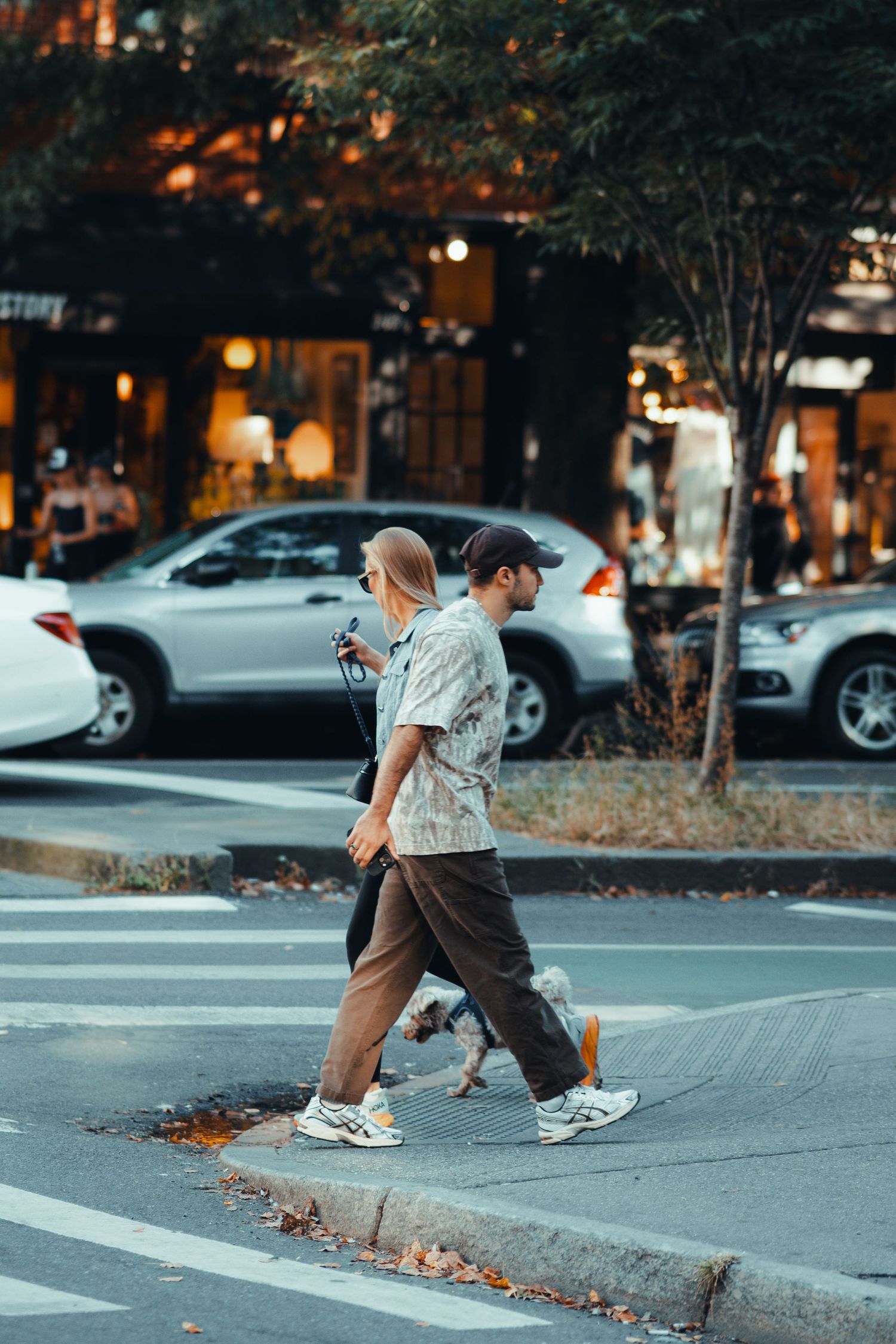 Two people crossing a street at dusk in front of a lit storefront and parked cars