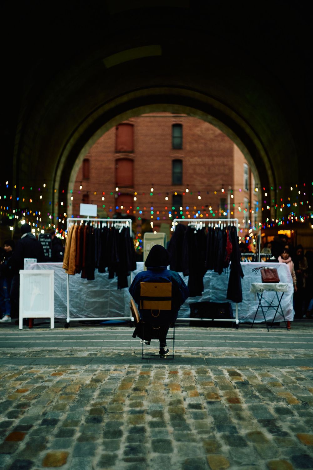 A vendor seated under a lit brick archway over cobblestones, market stalls set up beneath it