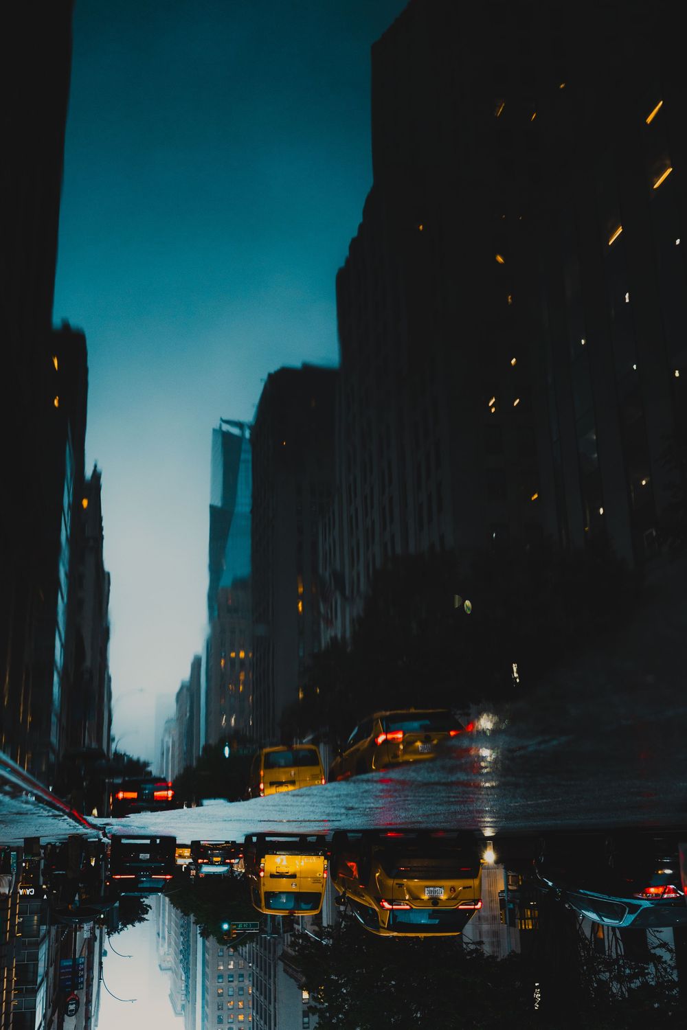 A wet avenue at night reflecting yellow cab lights, a strip of teal sky between the towers