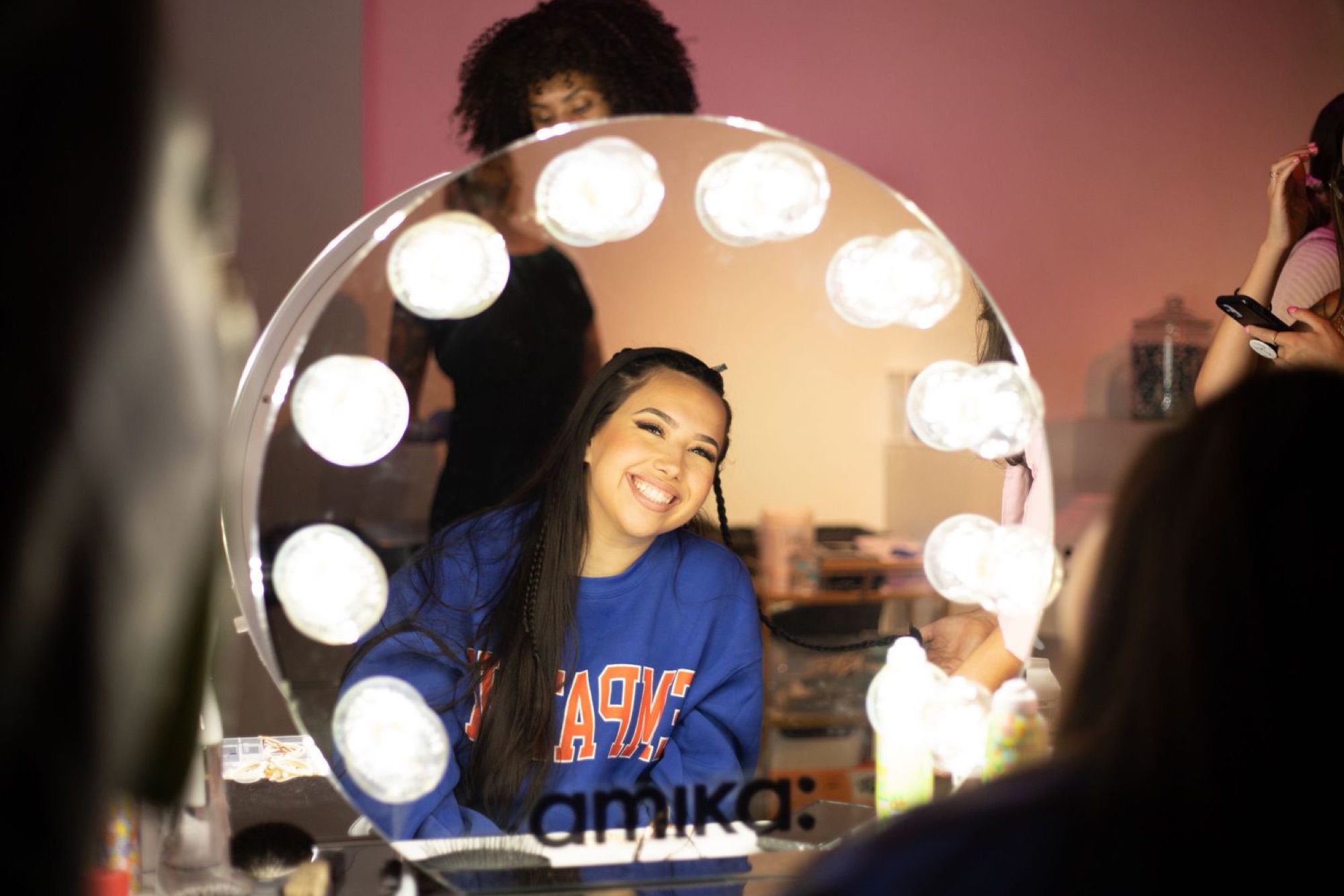 A model reflected in a round backstage vanity mirror lit by bulbs and etched with the amika logo, smiling as a hairstylist works behind her