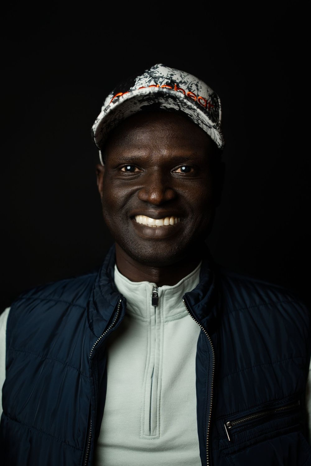 Studio portrait of a man in a navy vest over a white turtleneck and a beaded headband, lit against a black background