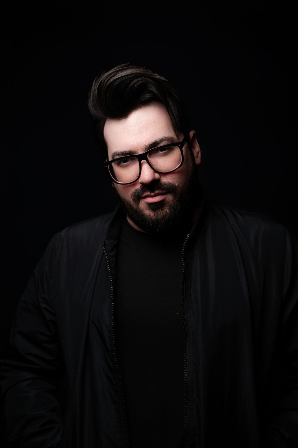 Studio portrait of a bearded man in glasses and a black jacket, lit against a black background