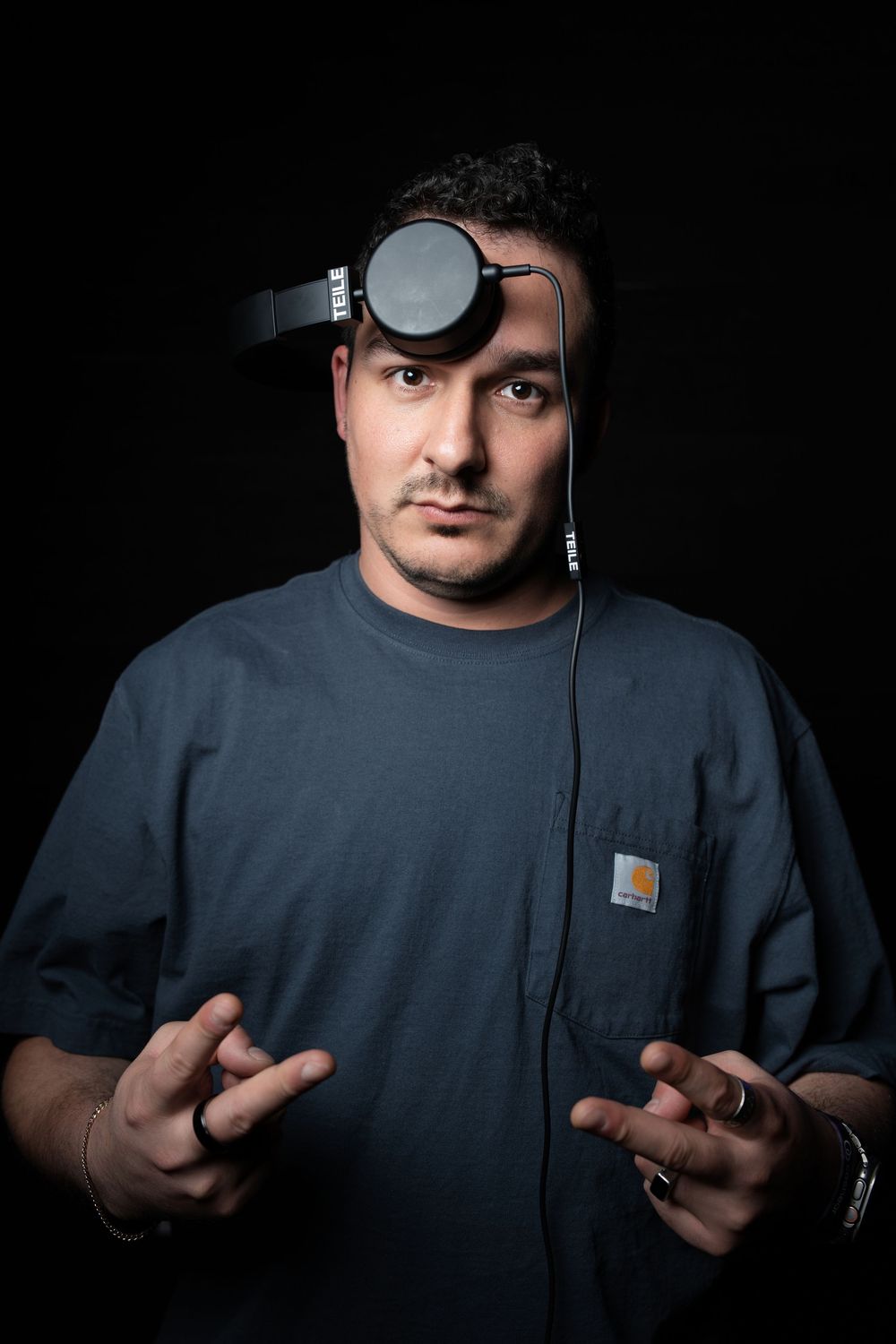 Studio portrait of a man in a grey tee with headphones pushed up onto his head, both hands raised during a gesture