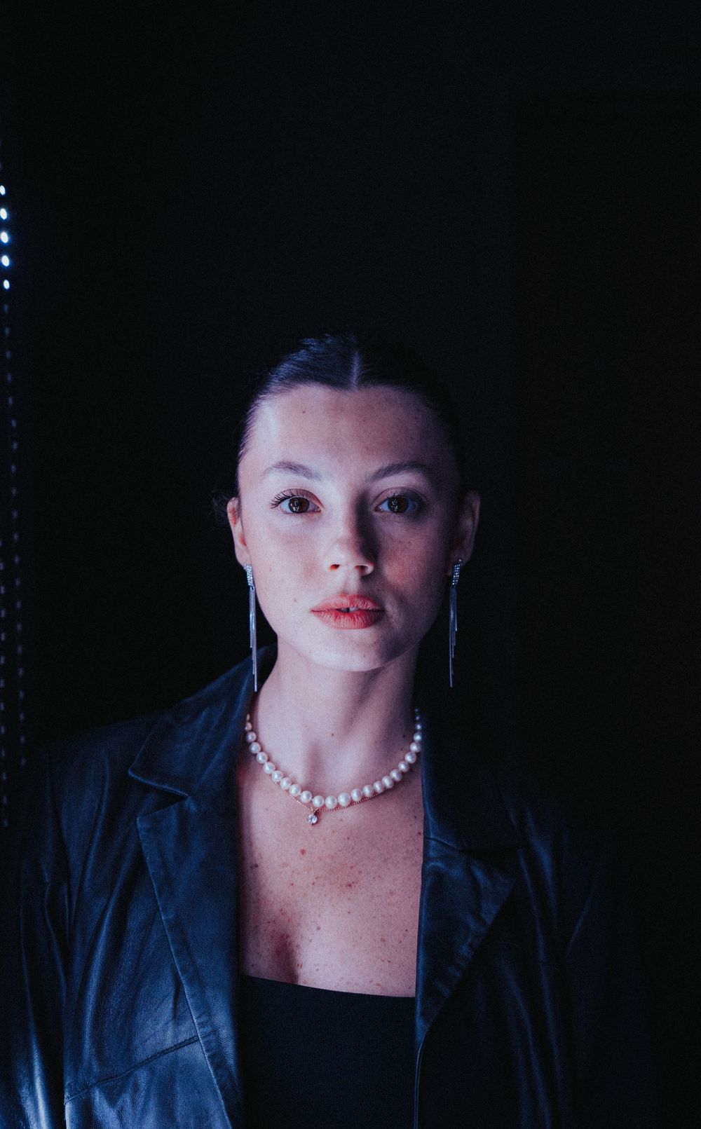 Night portrait of a woman in a black leather jacket and a pearl necklace, a lit sign thrown out of focus behind her