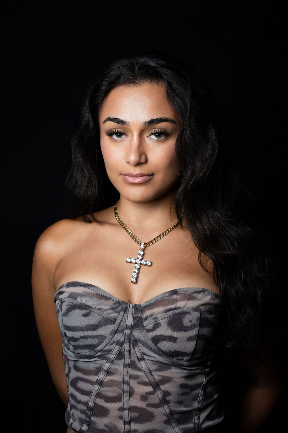 Studio portrait of a woman in a bandeau top with a leopard print and a cross pendant, lit against a black background