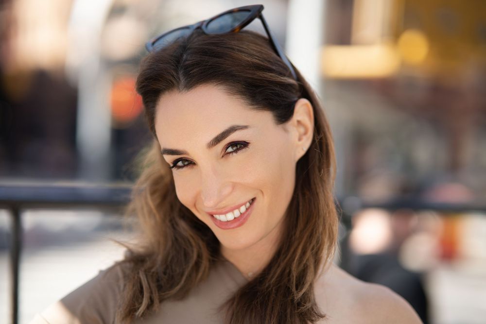 A woman with sunglasses pushed into her hair smiling on a city sidewalk, the street behind her thrown out of focus