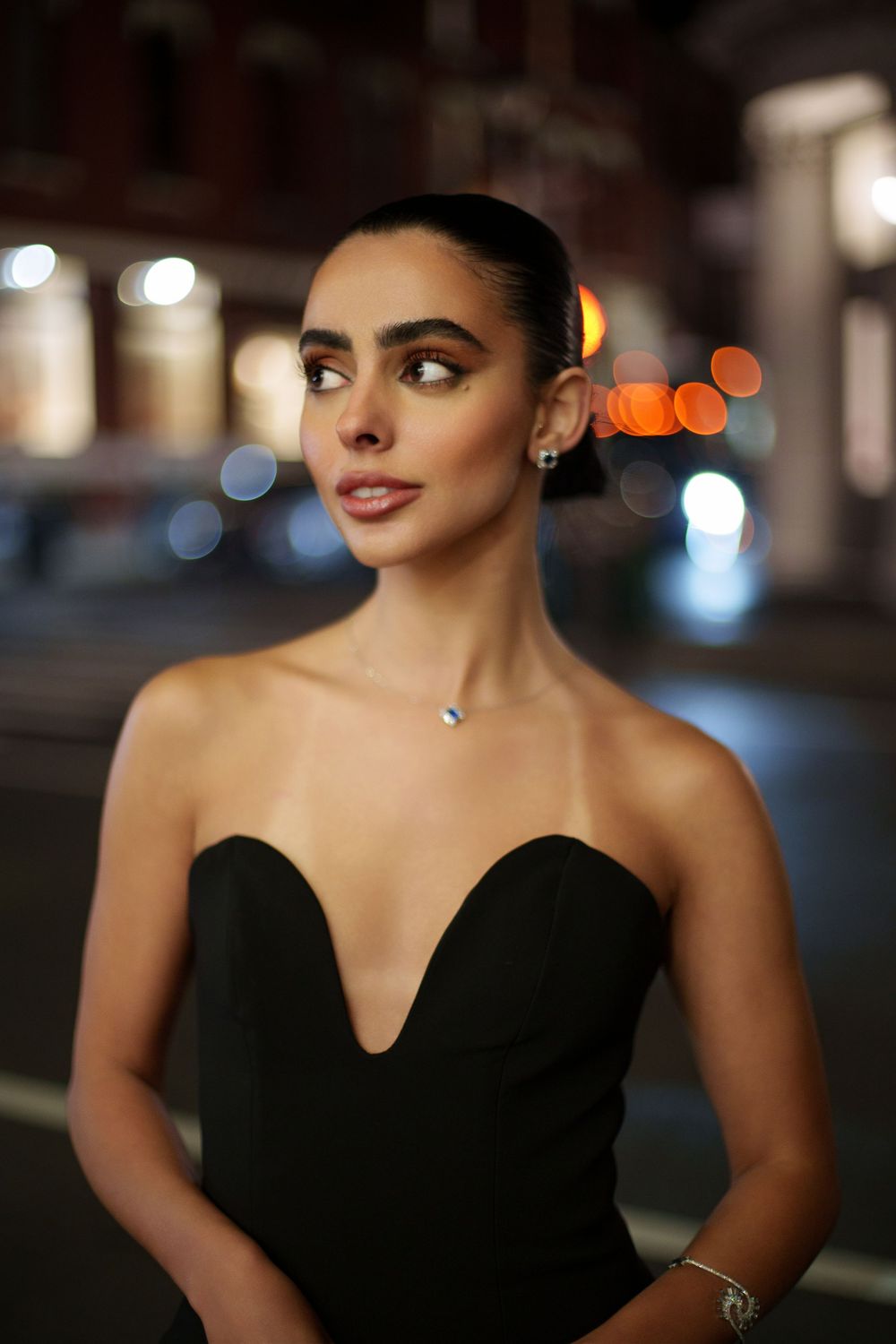 Night portrait of a model in a black strapless dress with her hair slicked back, street lights blurred behind her
