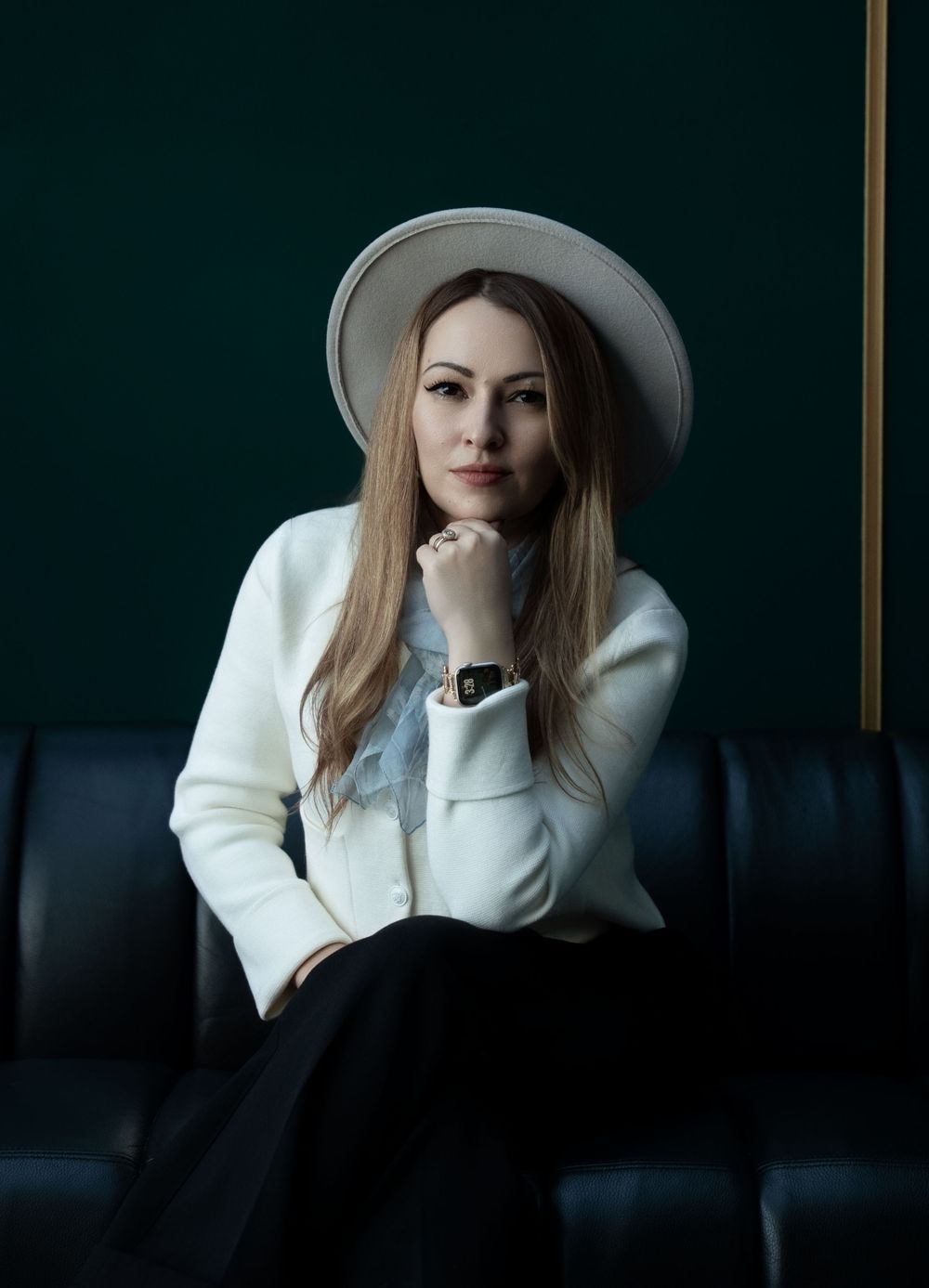 Model in a white coat and a pale hat with a wide brim, seated in a dark green leather booth