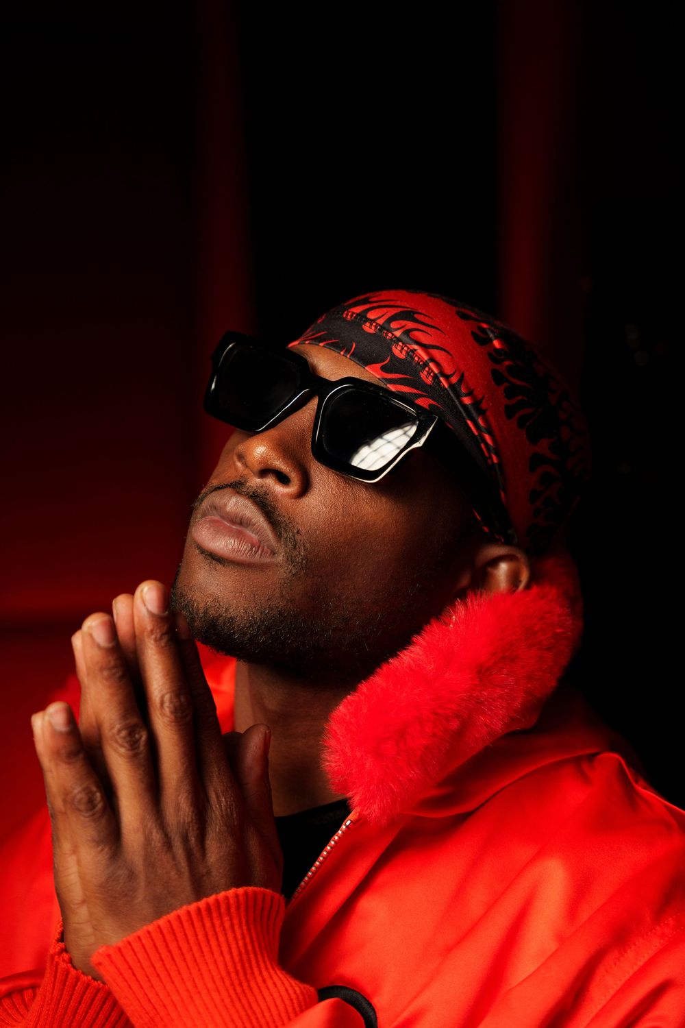 Portrait of a man in a red bandana, dark sunglasses and a jacket trimmed with red fur, hands pressed together under red light