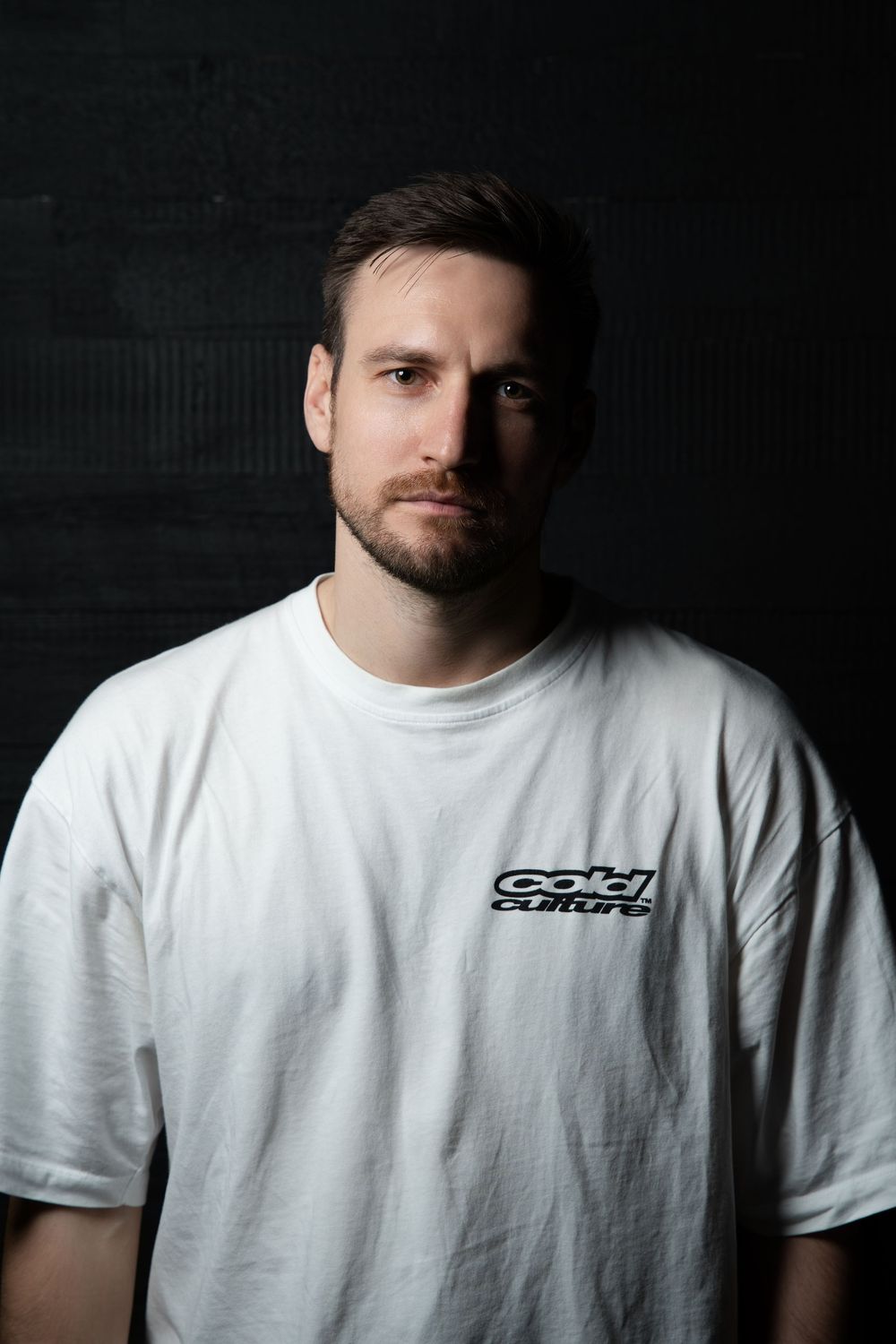 Studio portrait of a bearded man in a white printed tee, lit against a black background