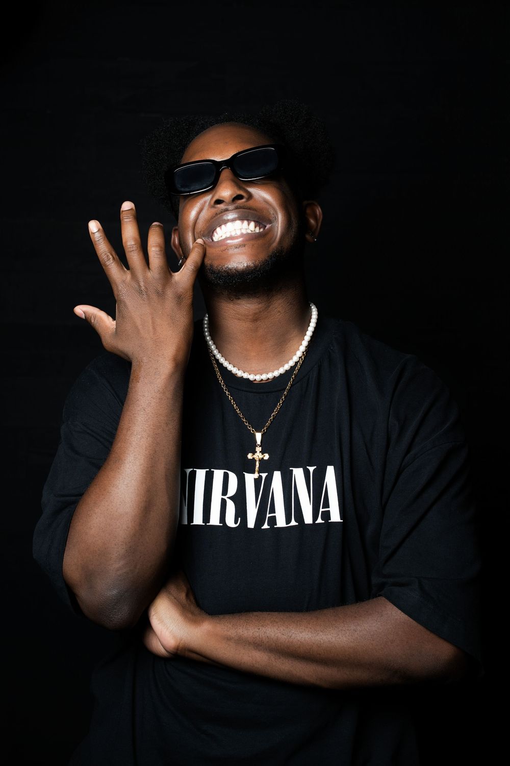Studio portrait of a man in sunglasses and a gold chain wearing a band tee, one hand raised beside his face