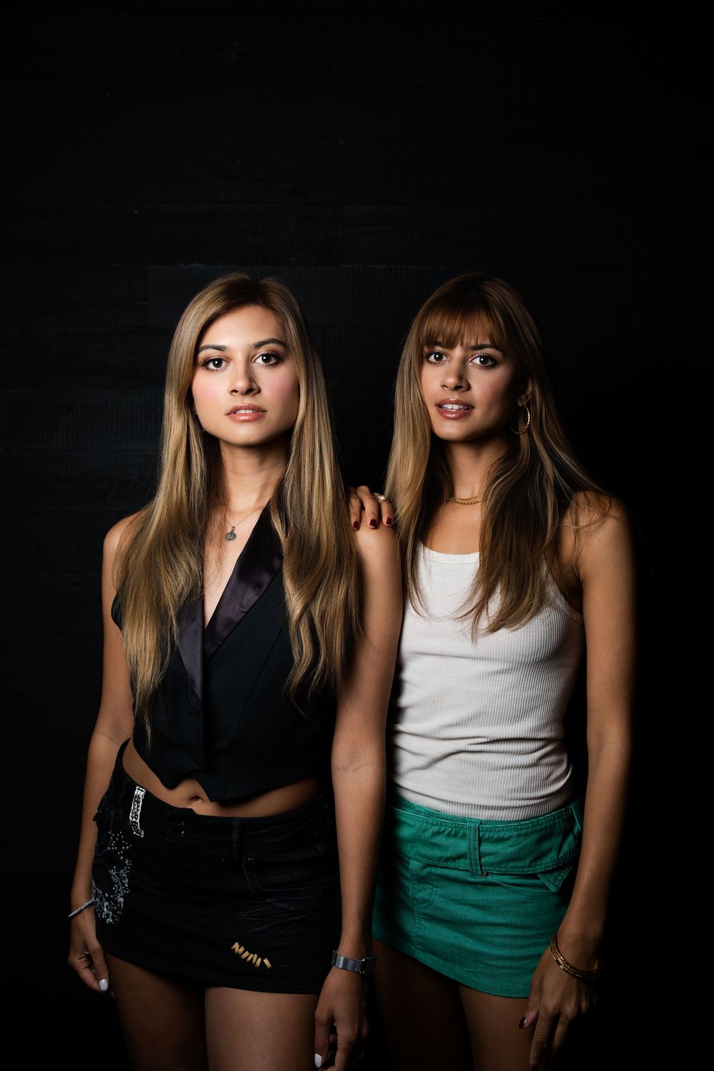 Two models in a black slip and a white tank with a green skirt, standing together against a black studio wall