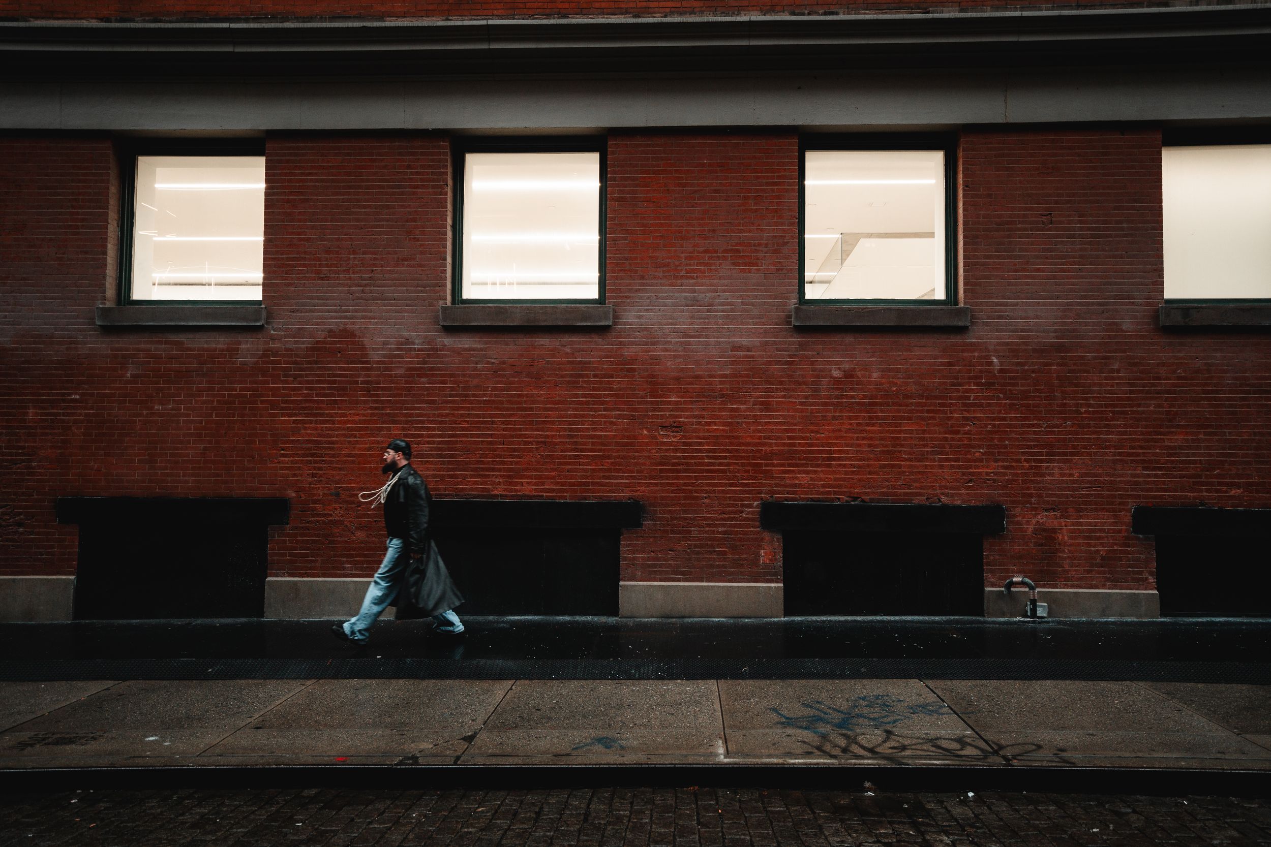 A person in a denim jacket walking past a long red brick wall lined with lit windows