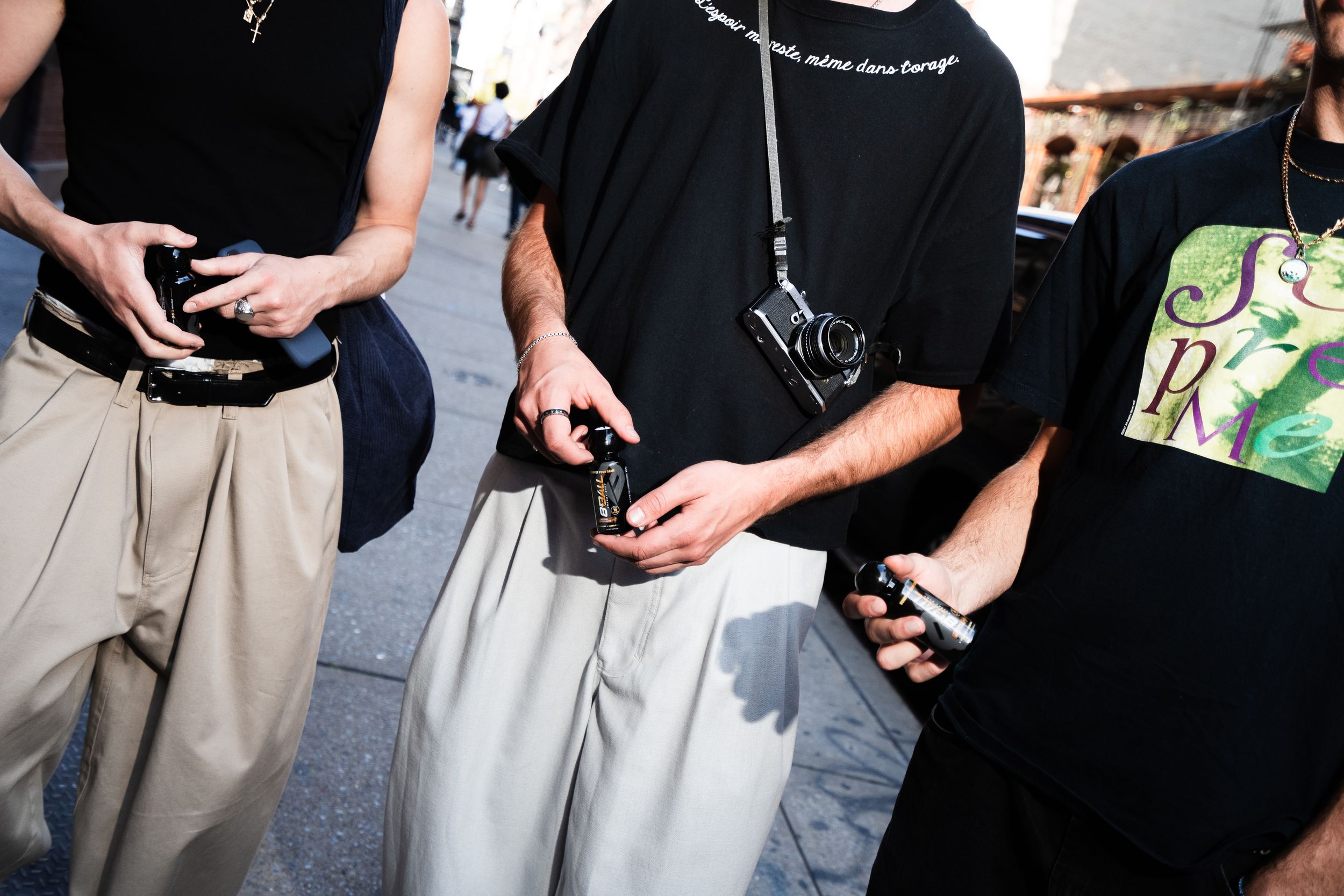 Three men walk a New York sidewalk in black tees and pleated light trousers, one with a film camera around his neck, each holding a small black energy shot bottle
