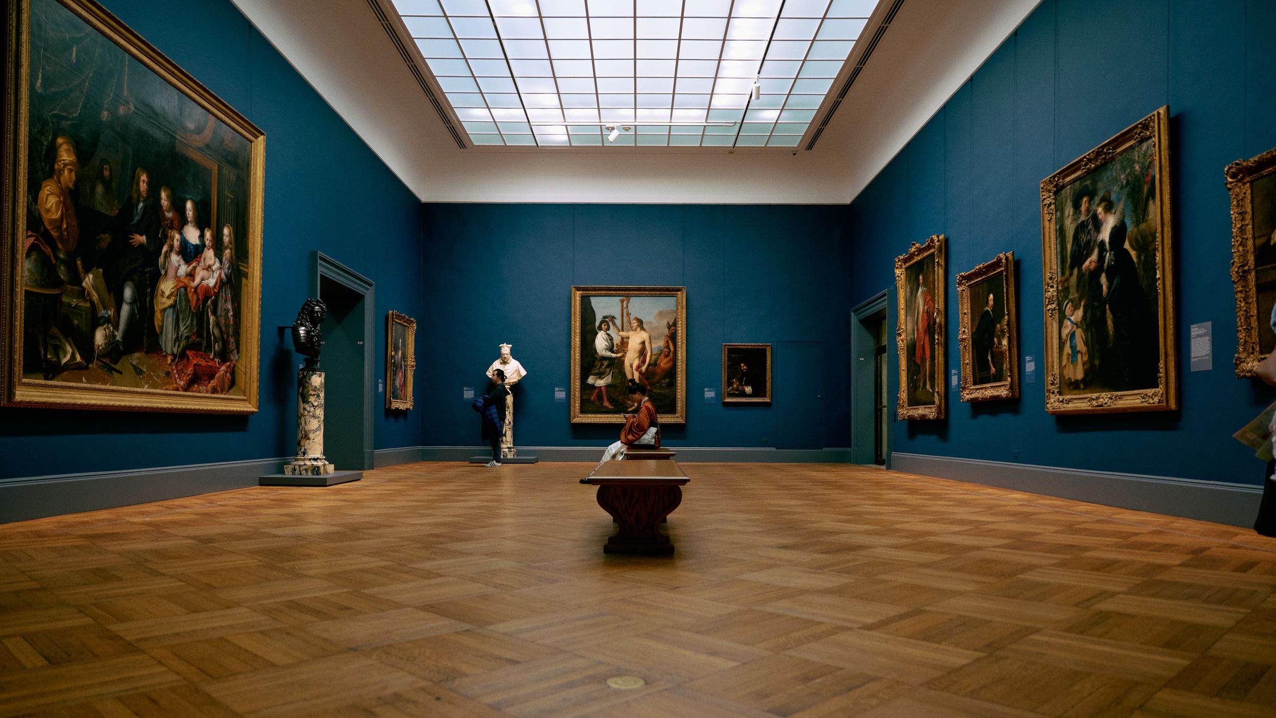 A wide blue museum gallery hung with paintings in gilt frames, a single sculpture at the centre of the floor