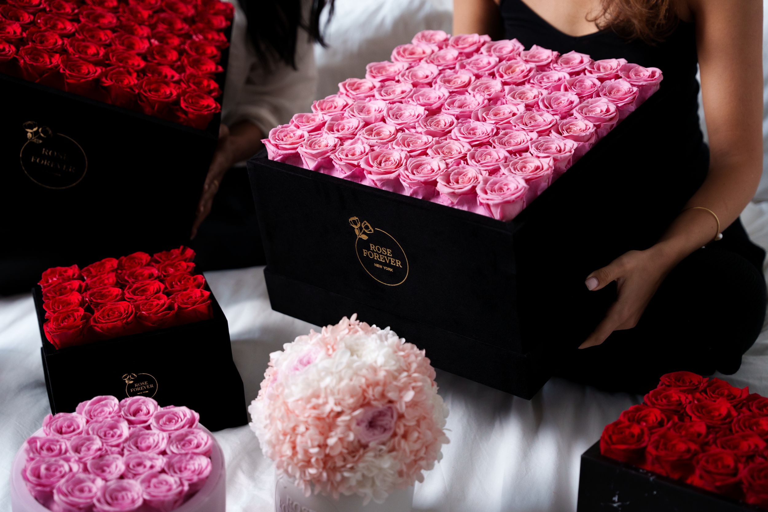 Two women with black velvet Rose Forever boxes of pink and red preserved roses on a bed