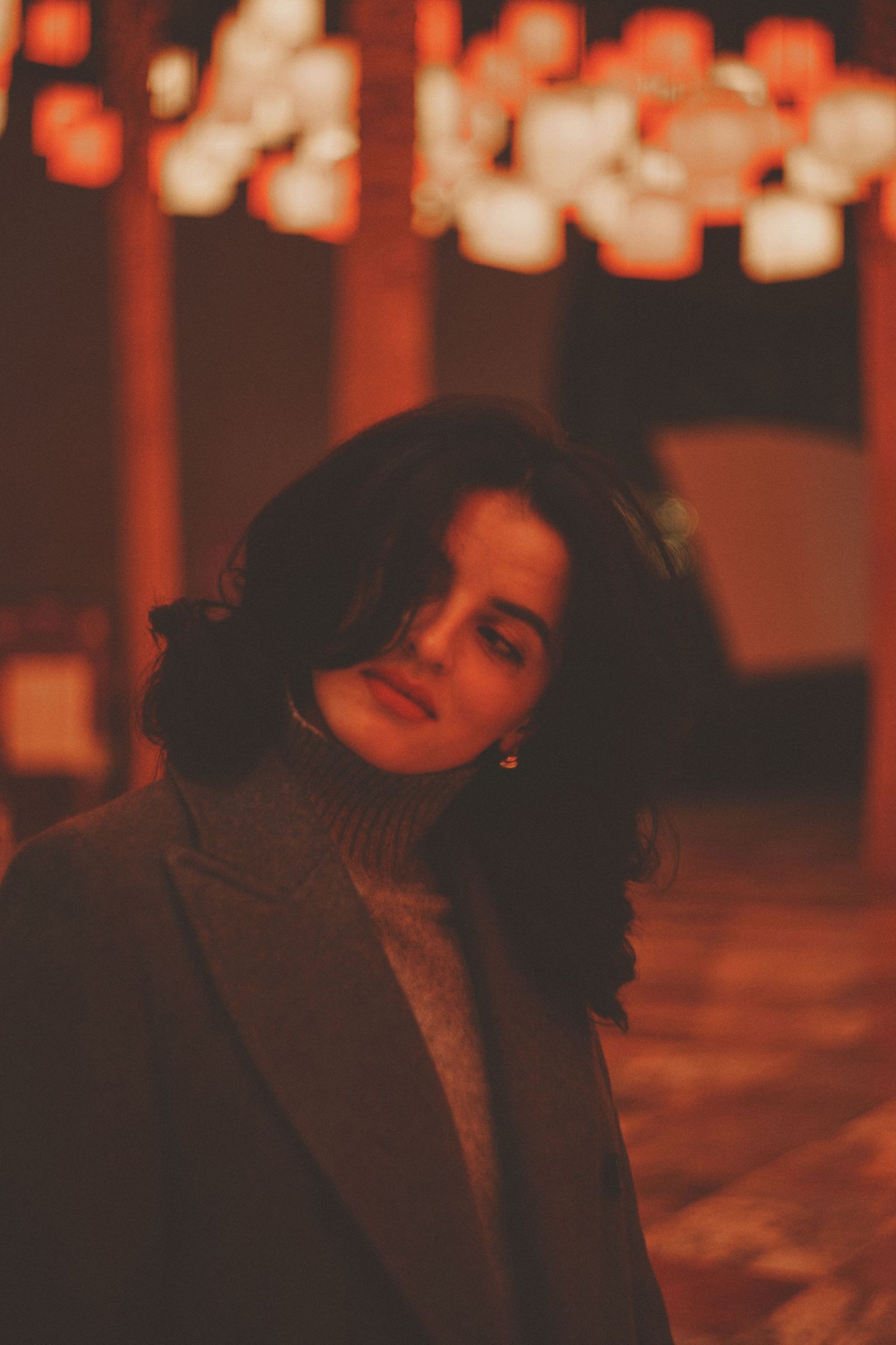 Portrait of a woman in a gray turtleneck and coat looking down softly, framed by warm blurred lanterns at night