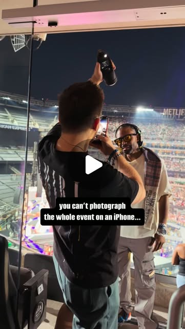 Filming a skit in a stadium suite: one man photographs another holding a drink over the lit field at MetLife