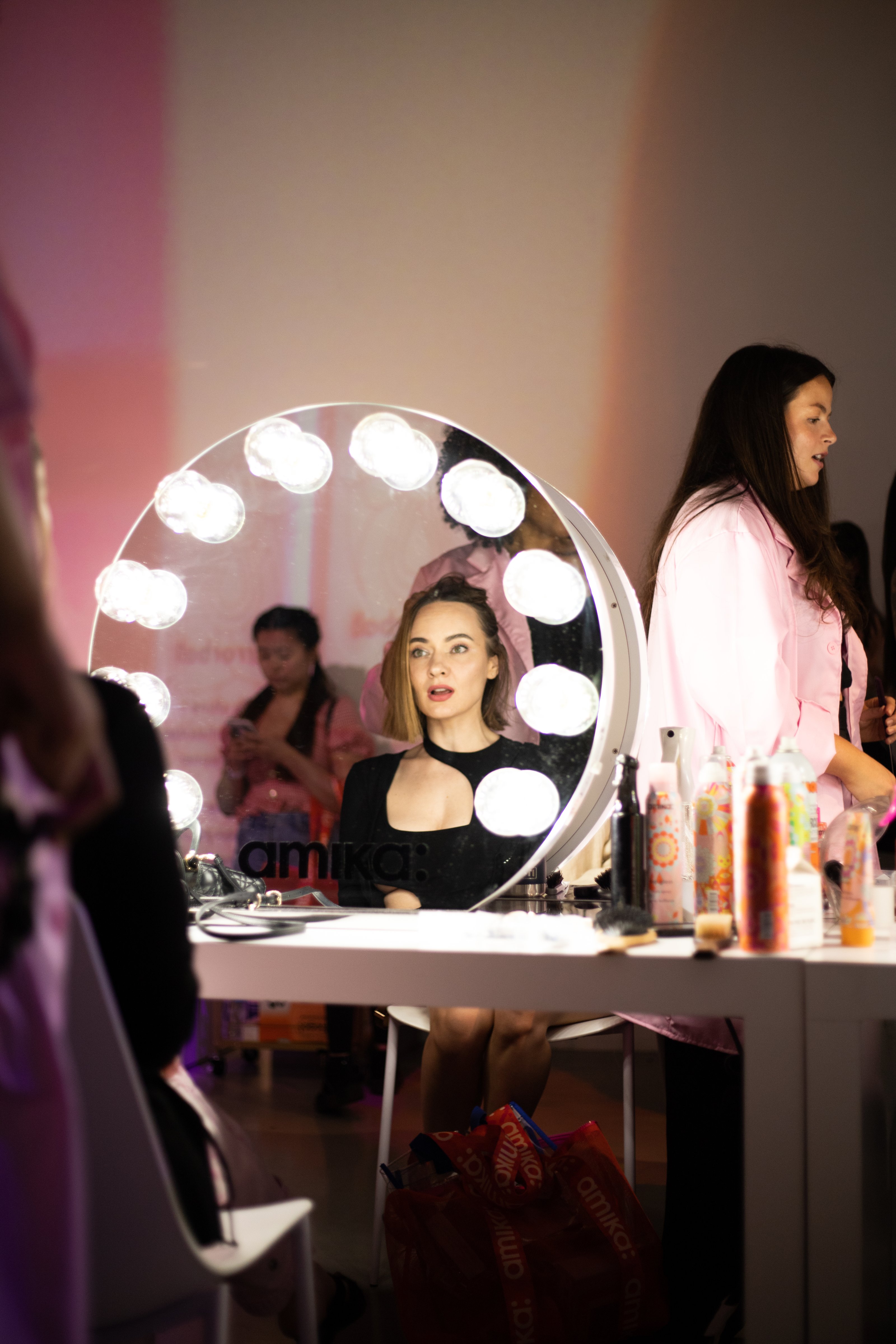 A woman in a black top looks ahead from inside a round lighted vanity mirror during the Amika event at Lume Studios in Tribeca, Manhattan.