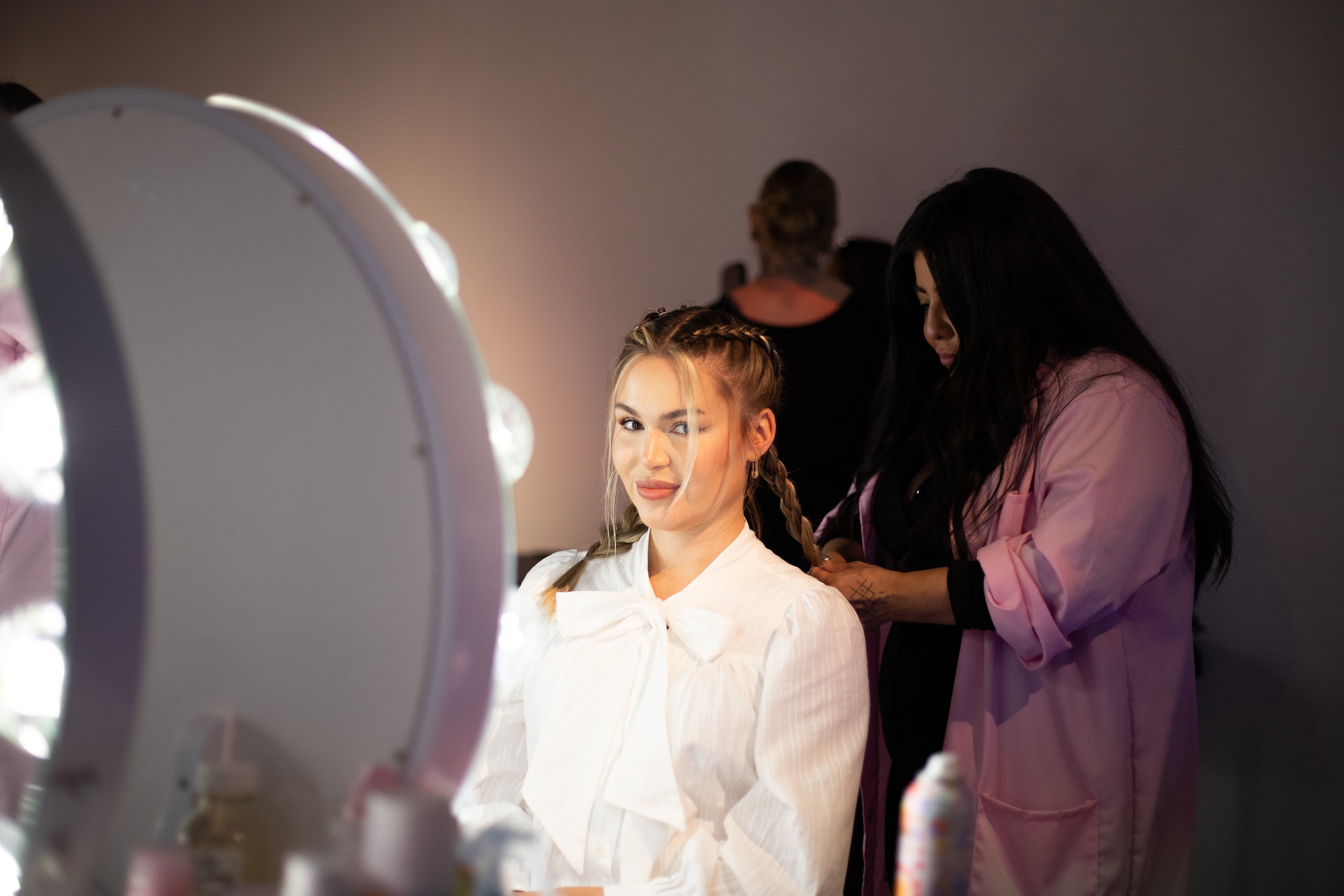 A woman in a white blouse smiles as a stylist braids her hair before a lighted vanity for Amika at Lume Studios in Tribeca, Manhattan.
