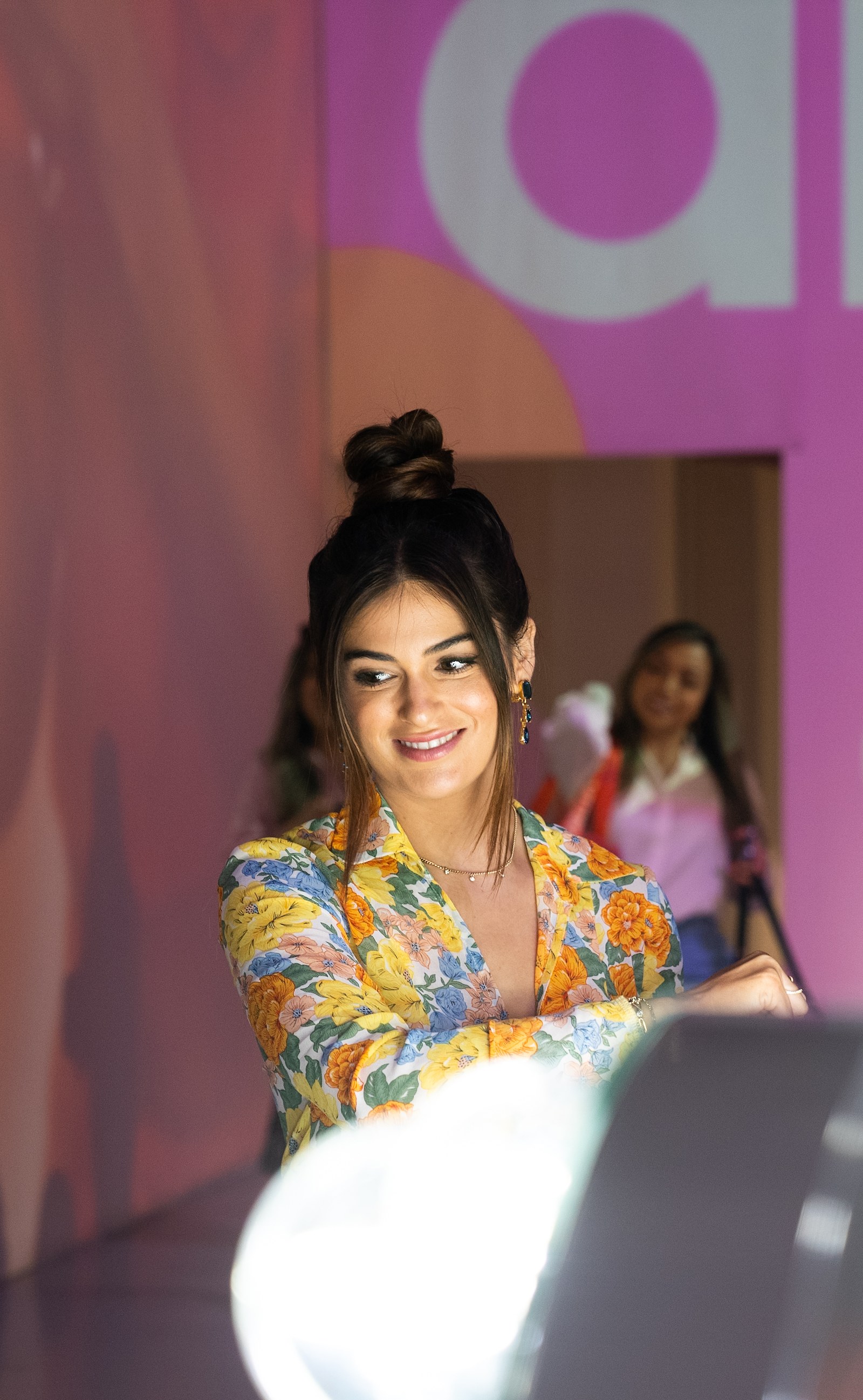 A woman with a high hair bun and floral blouse smiles beside a lighted mirror for Amika at Lume Studios in Tribeca, Manhattan.