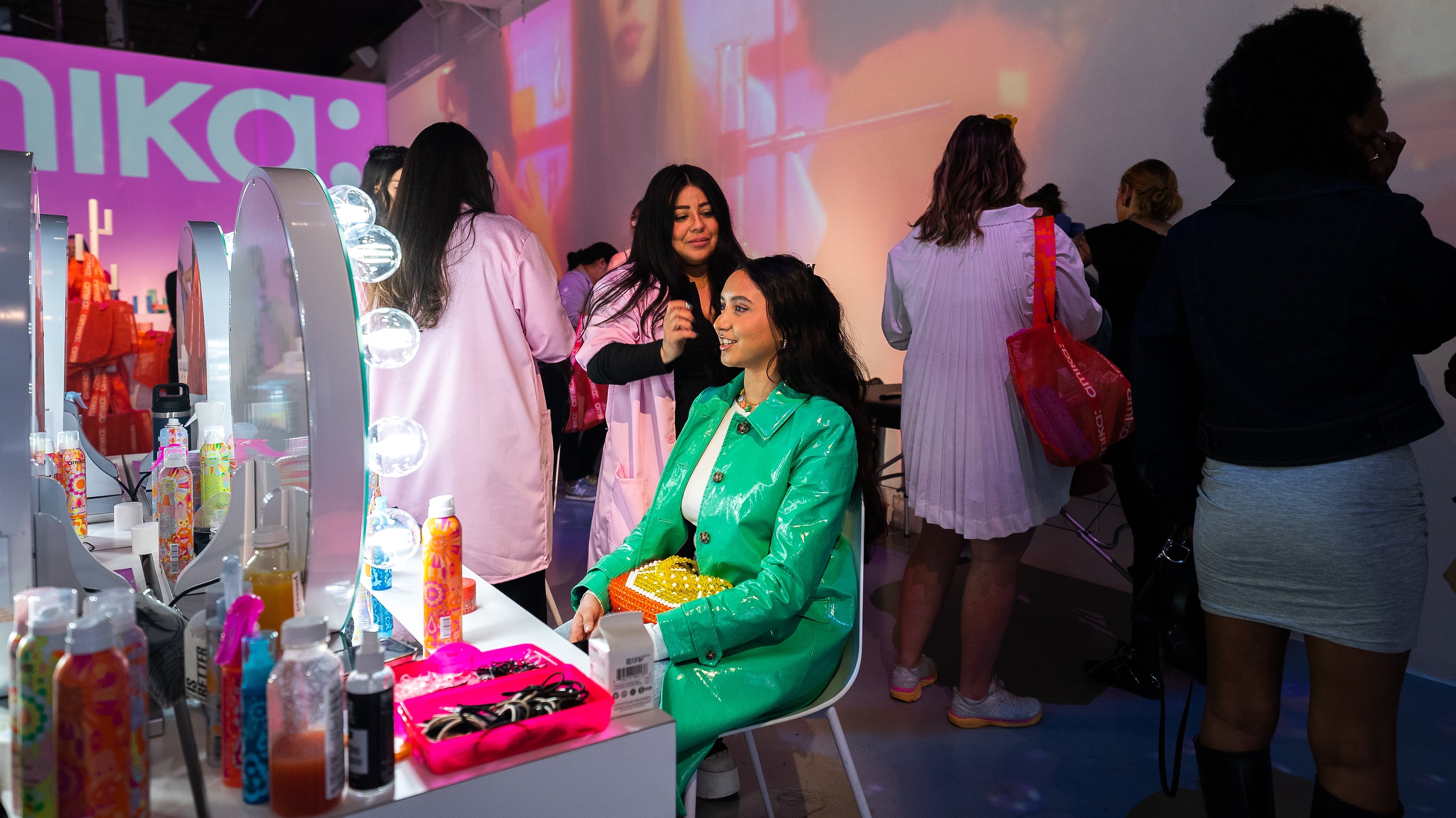 A woman in a green coat smiles while getting her hair styled at an illuminated vanity for Amika at Lume Studios in Tribeca, Manhattan.