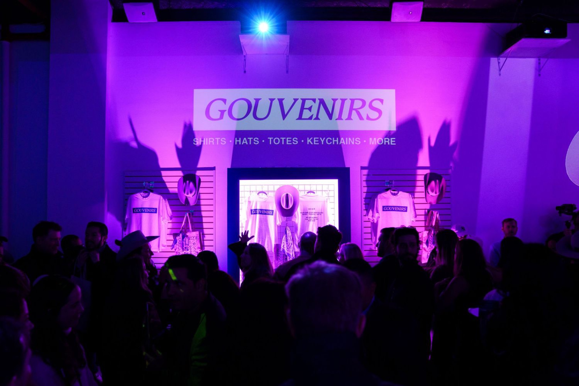 Guests inside the Gouvenirs shop under purple projected branding, with racks of branded merchandise on the walls