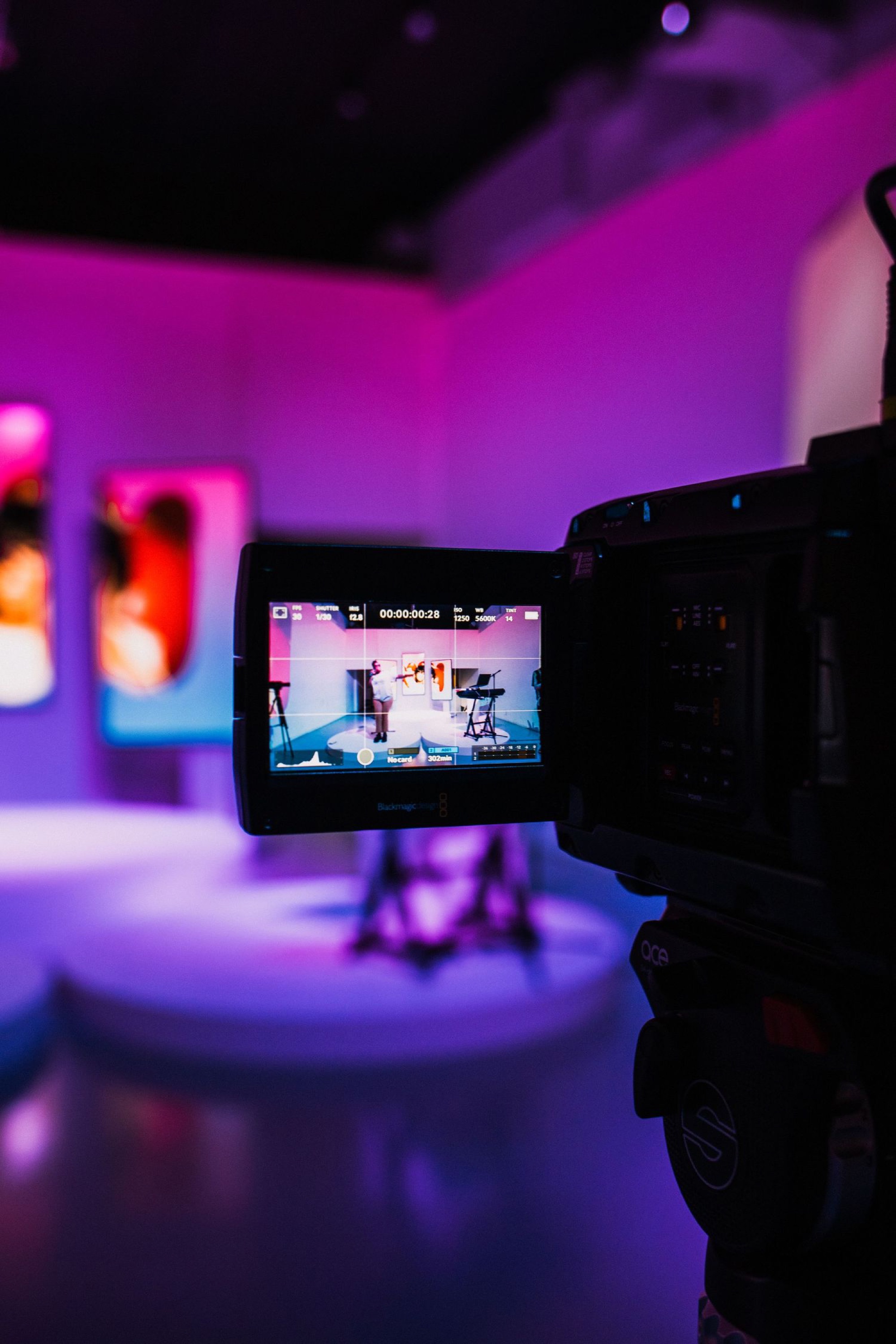 The hinged LCD monitor on a cinema camera shows the framed shot of a performer in a studio set lit in purple and pink, with other cameras and gear visible around it