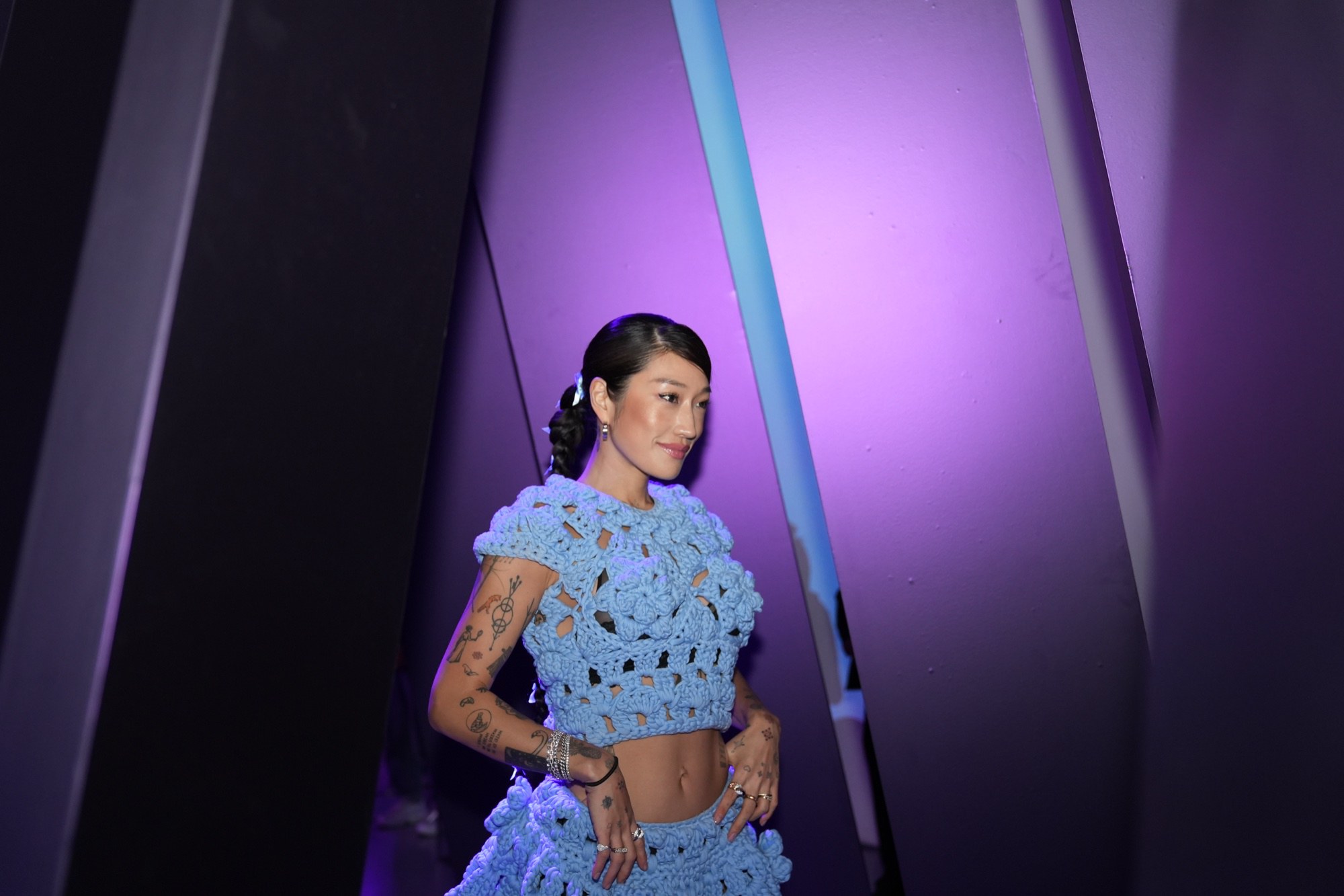 A woman in a blue crochet top and skirt walks between tall angled panels under purple and blue light
