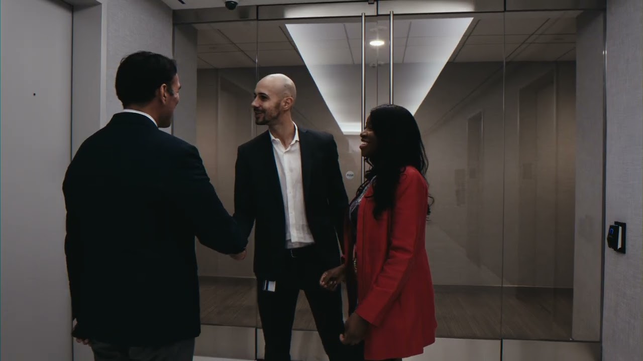 Frame from the Ferrari Express New York film: two people in suits shake hands while a woman in a red blazer smiles in an office lobby