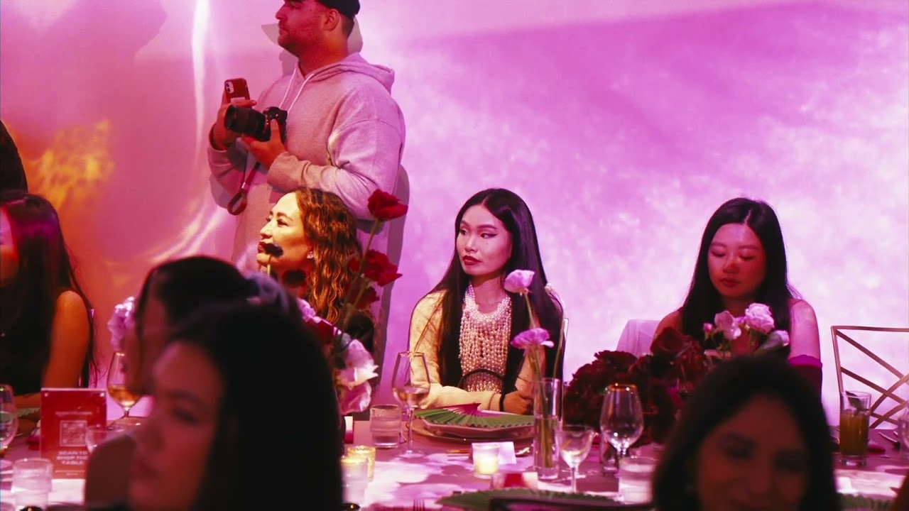 Guests seated at a flower lined dinner table washed in pink projection light as a photographer works behind them